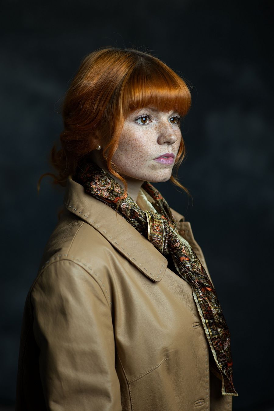 Portrait of a young redhead woman with freckles wearing a beige leather jacket and colorful scarf against dark background.