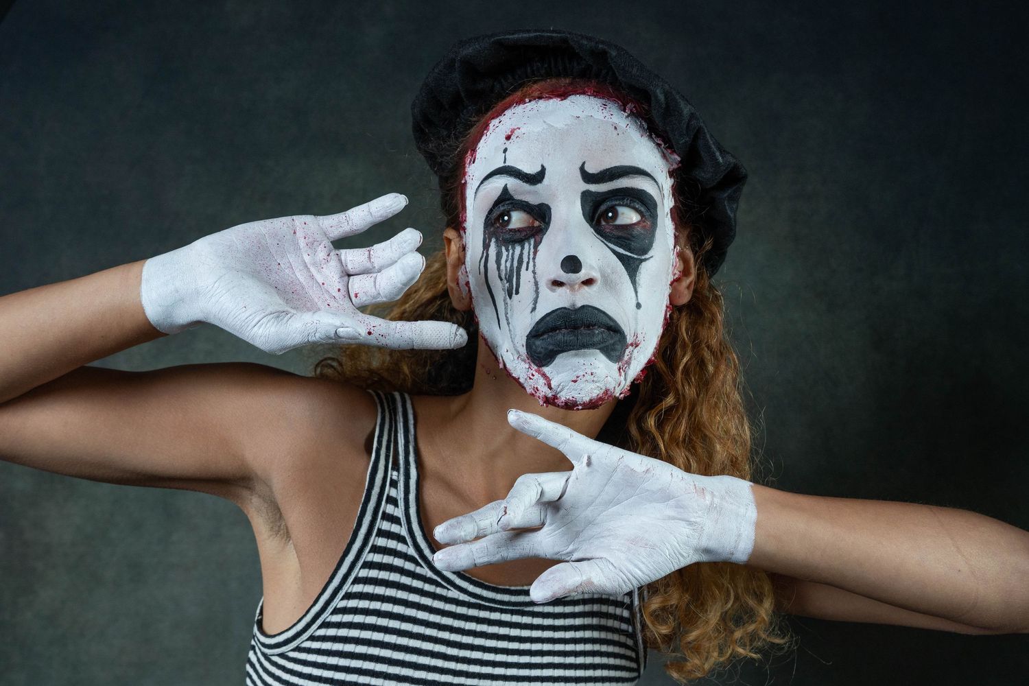 Woman in dramatic mime makeup with white face paint, black tears, and white gloves posing against dark background.