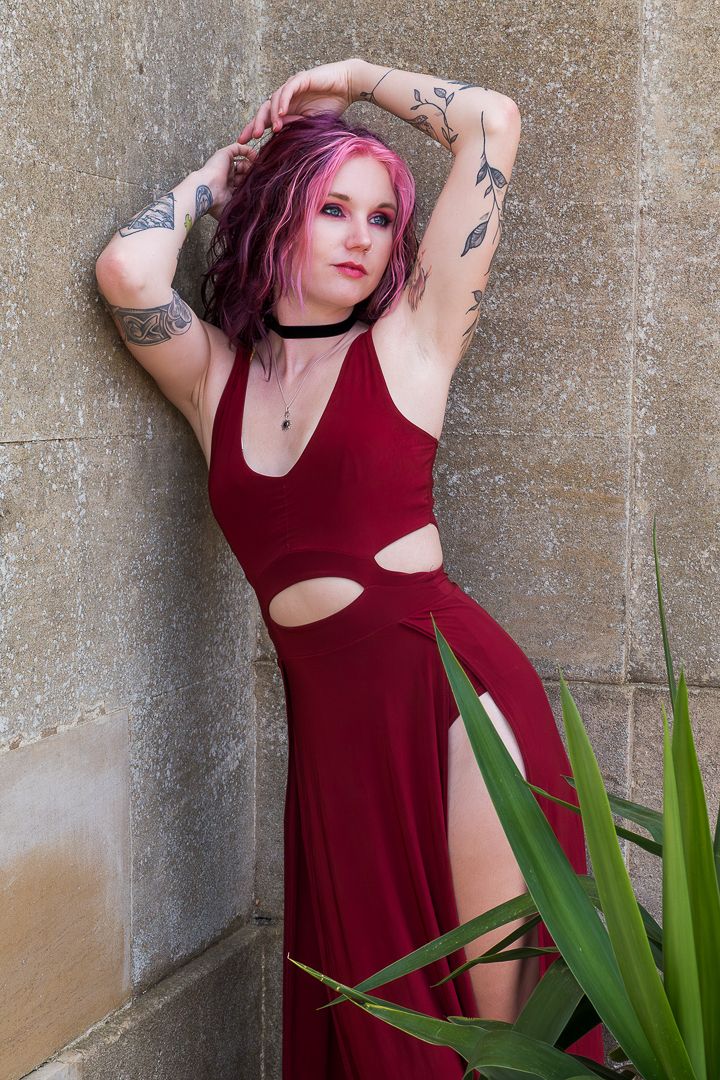 Woman with pink hair and tattoos posing in red cutout dress against stone wall with tropical plant.