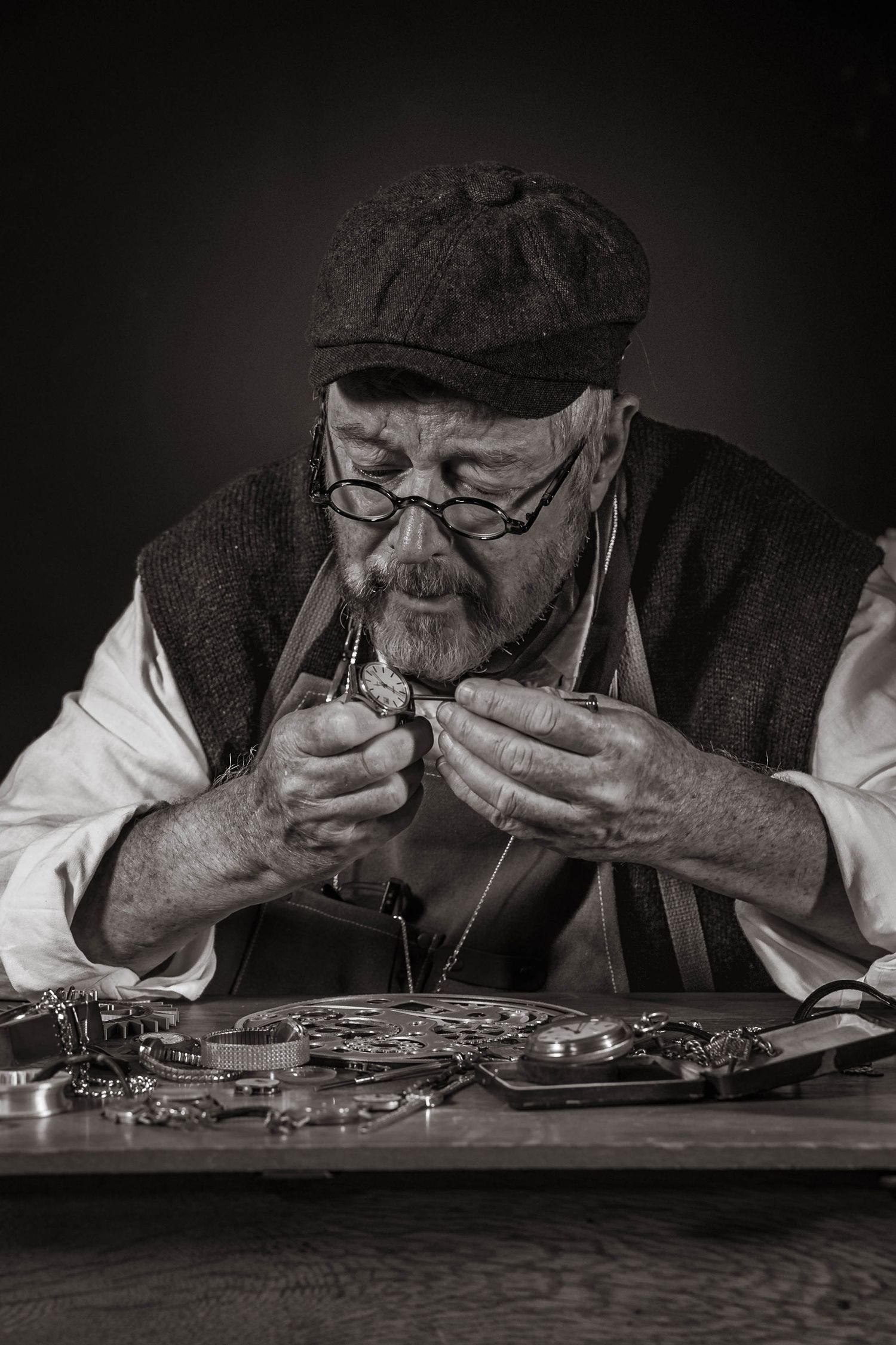 Elderly watchmaker in flat cap and glasses examines pocket watch at workbench in black and white photo.