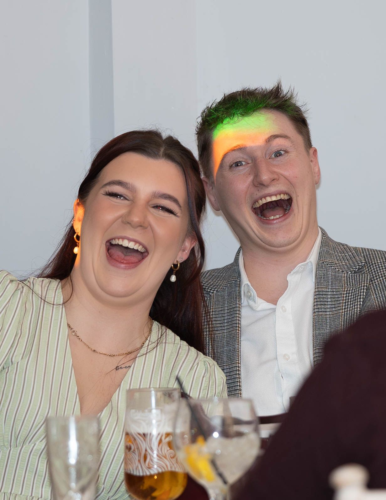 Two laughing friends at a social event, sharing a joyful moment together at a table with drinks.