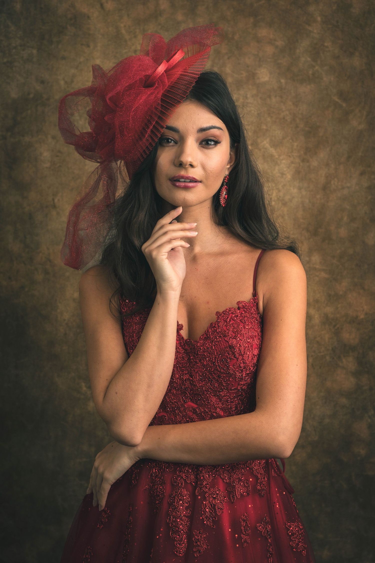 Woman in red lace gown and matching fascinator hat poses elegantly against a vintage brown backdrop.