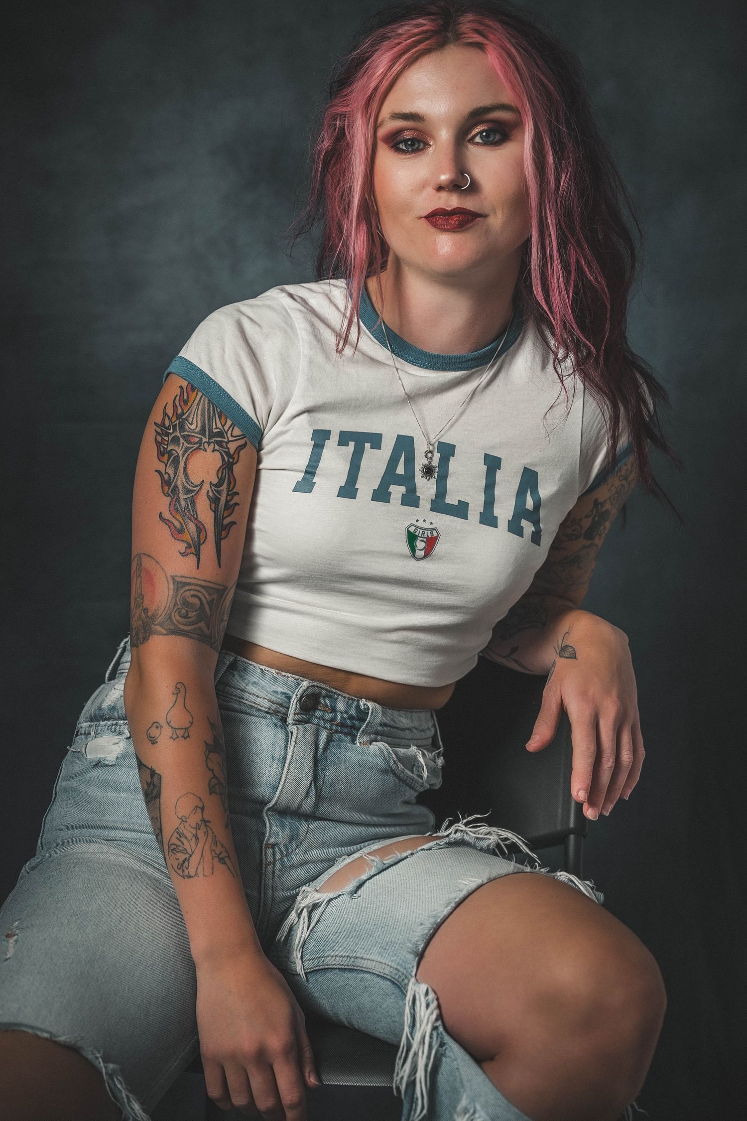 Tattooed woman with pink hair wearing Italia crop top and ripped denim shorts seated against dark background.