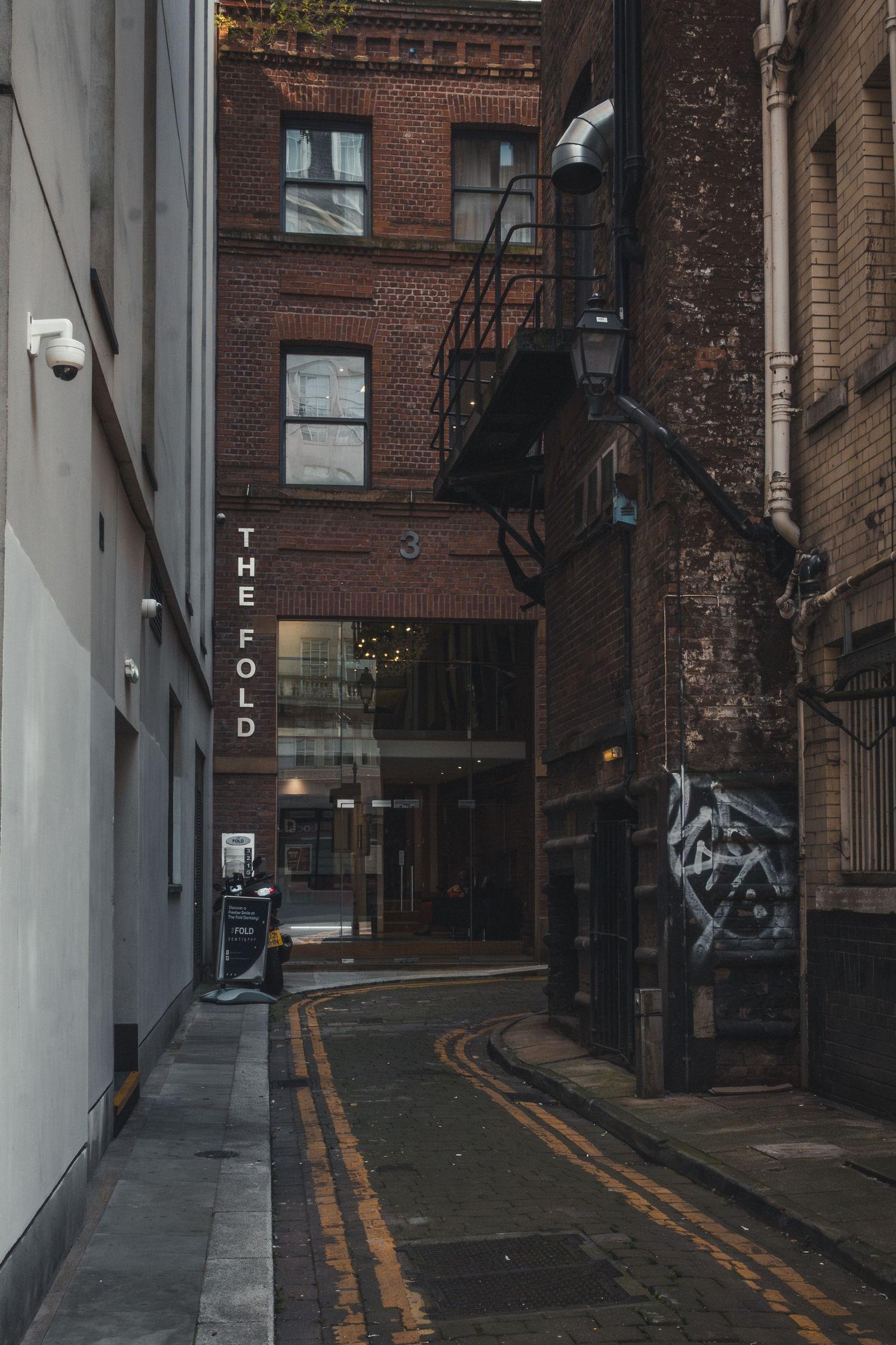 Narrow urban alley with brick buildings and double yellow lines painted on the ground between tall red walls.