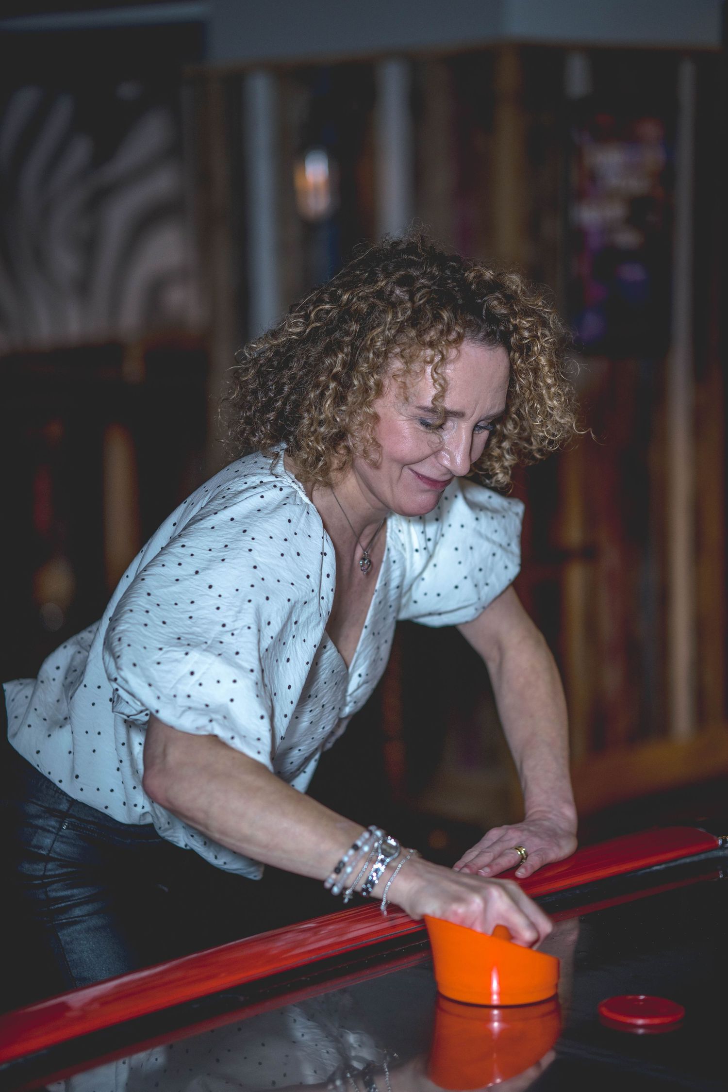 Woman with curly hair playing air hockey, leaning over red table holding orange mallet in arcade setting.