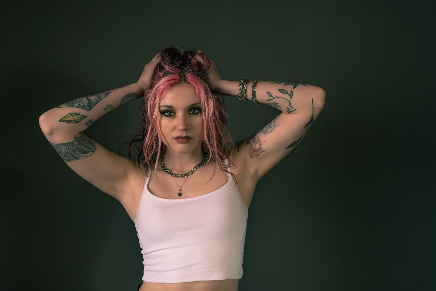 Young woman with pink hair and tattoos poses in white crop top against dark green background.