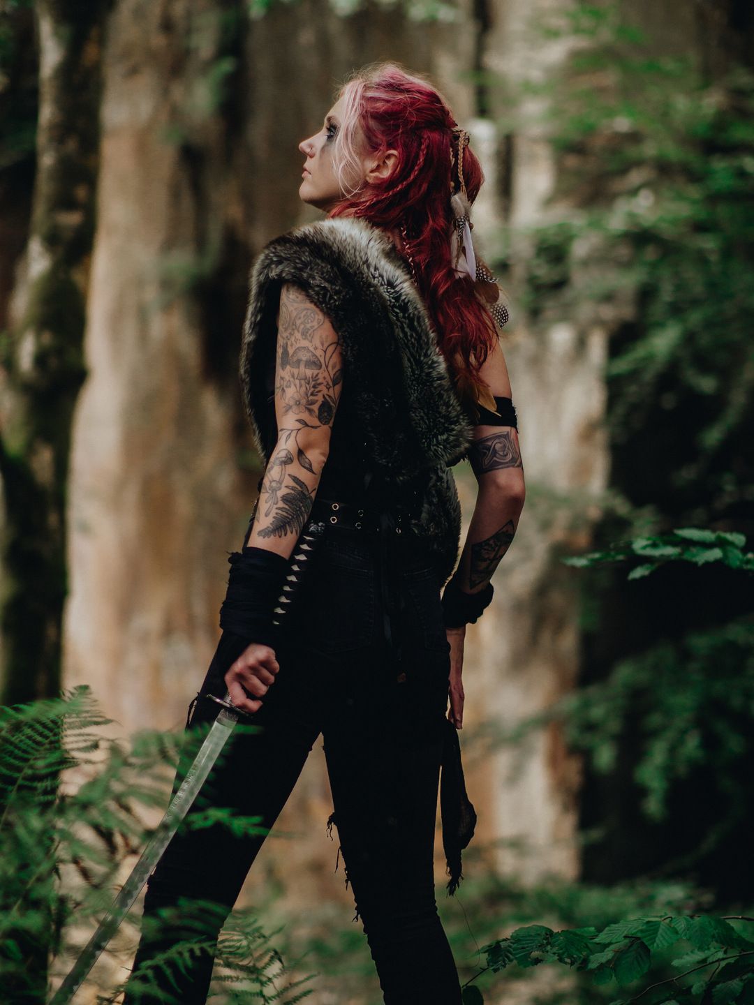 Tattooed woman with red hair in Viking warrior costume holding a dagger in a lush forest setting.