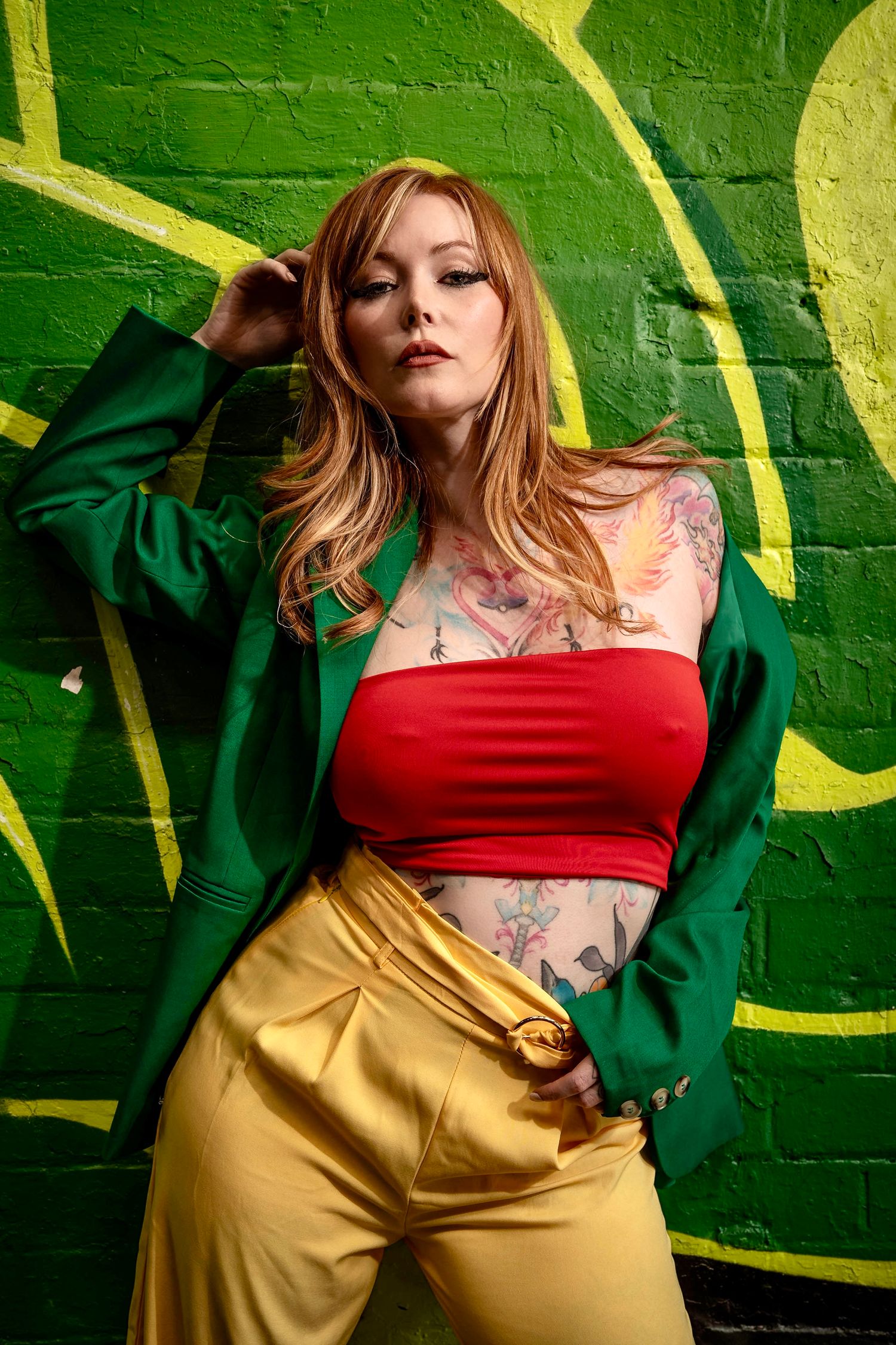 Tattooed woman in green blazer, red crop top, and gold pants posing against a vibrant green graffiti brick wall.