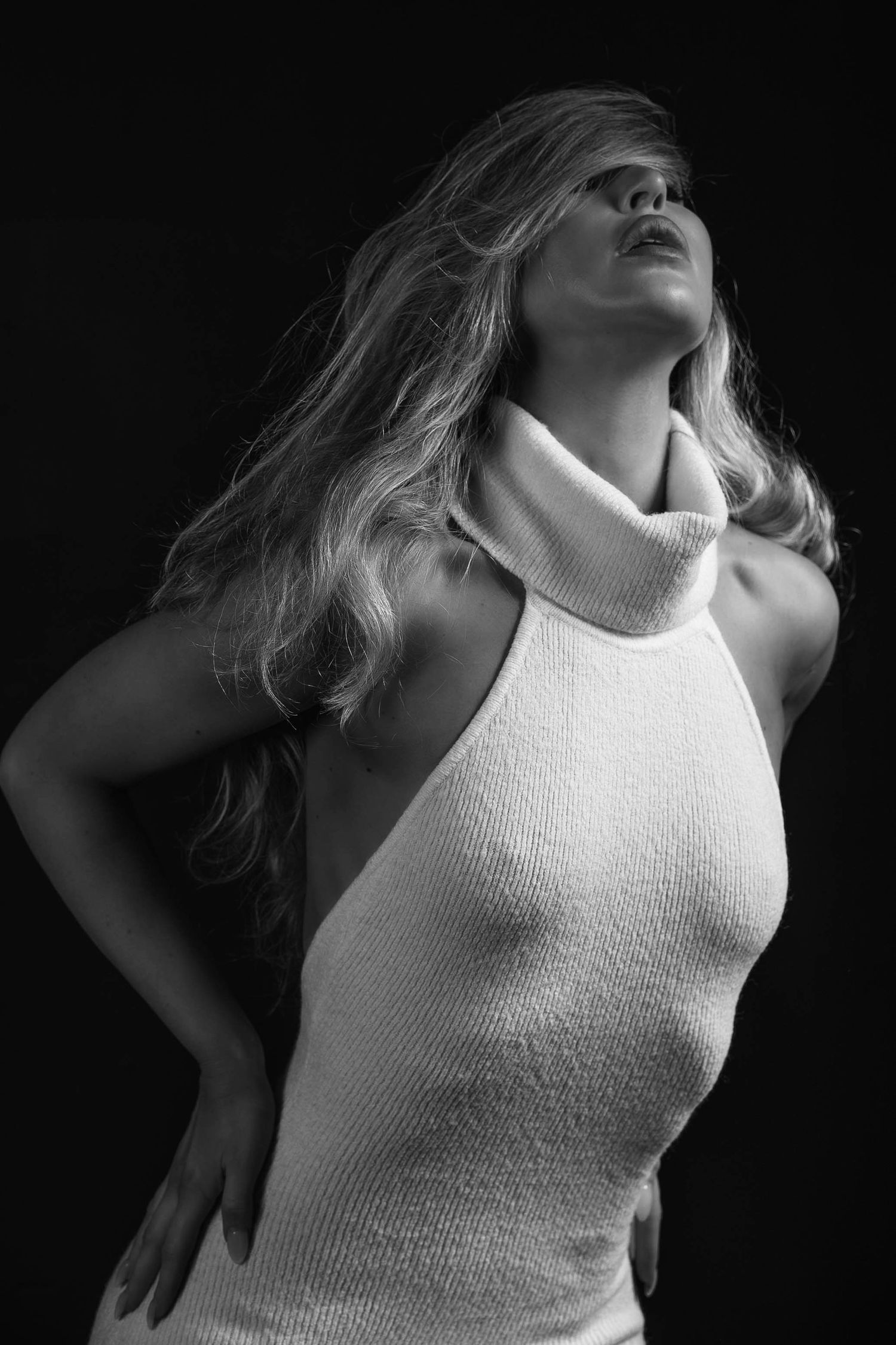 Black and white portrait of a blonde woman in a white turtleneck halter top posing against a dark background.