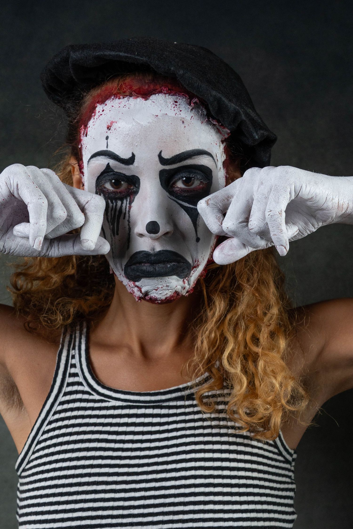 Creepy mime clown with white face paint, black tears, red hair, beret hat, striped shirt, raising gloved hands in scary pose.