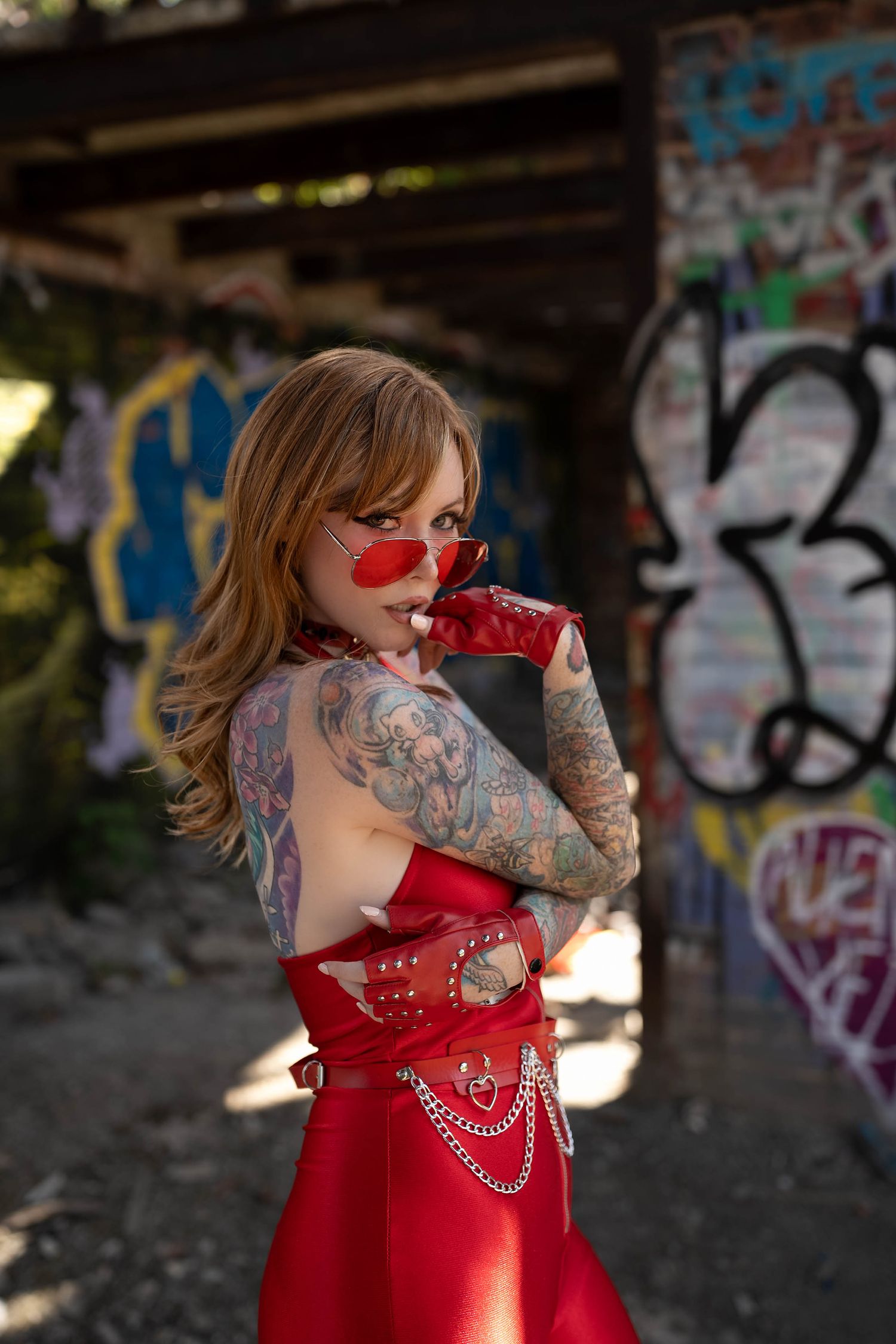 Tattooed woman in red backless dress and sunglasses poses before graffiti wall in urban setting.