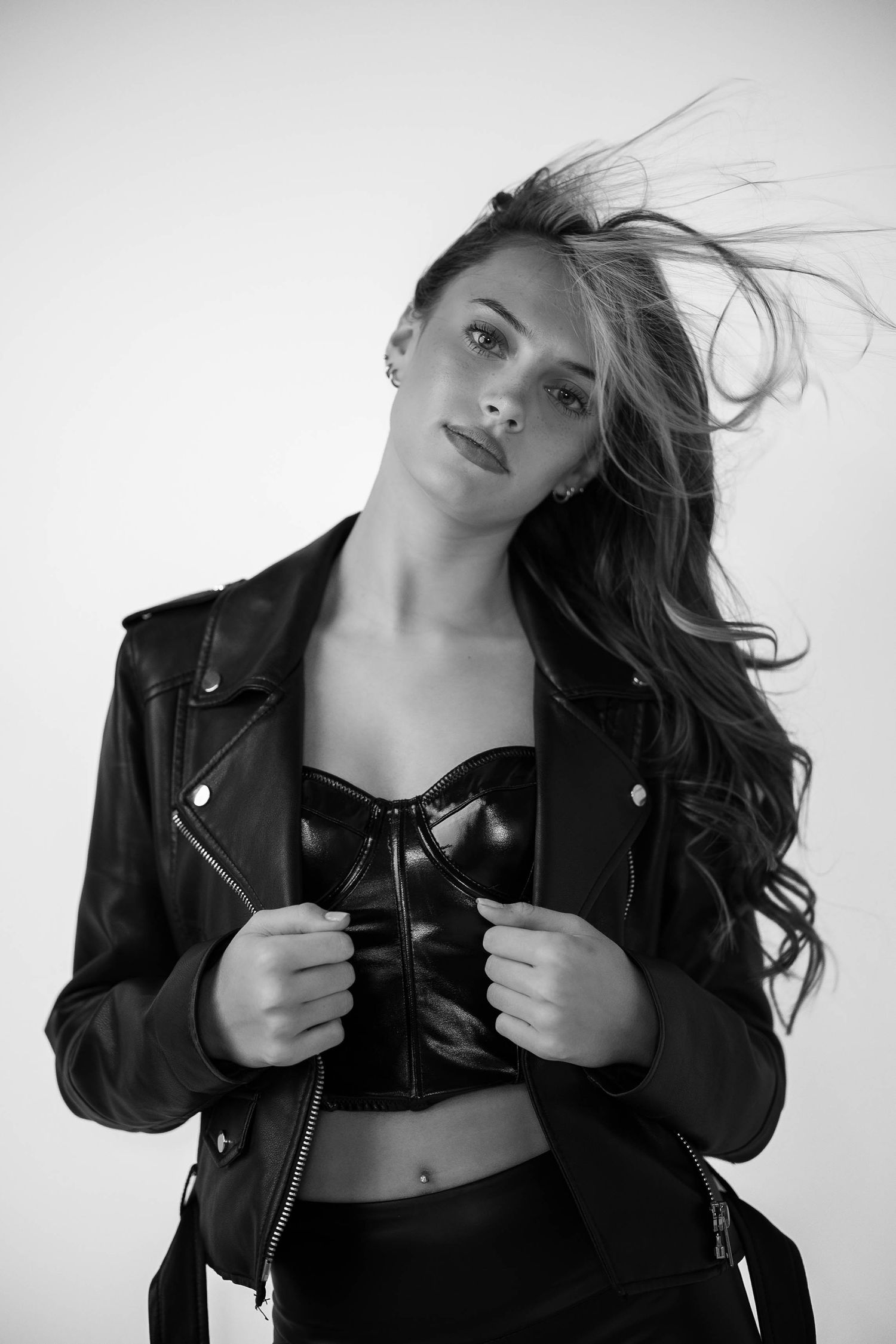 Black and white photo of a woman in leather jacket and bustier top with flowing hair against white background.