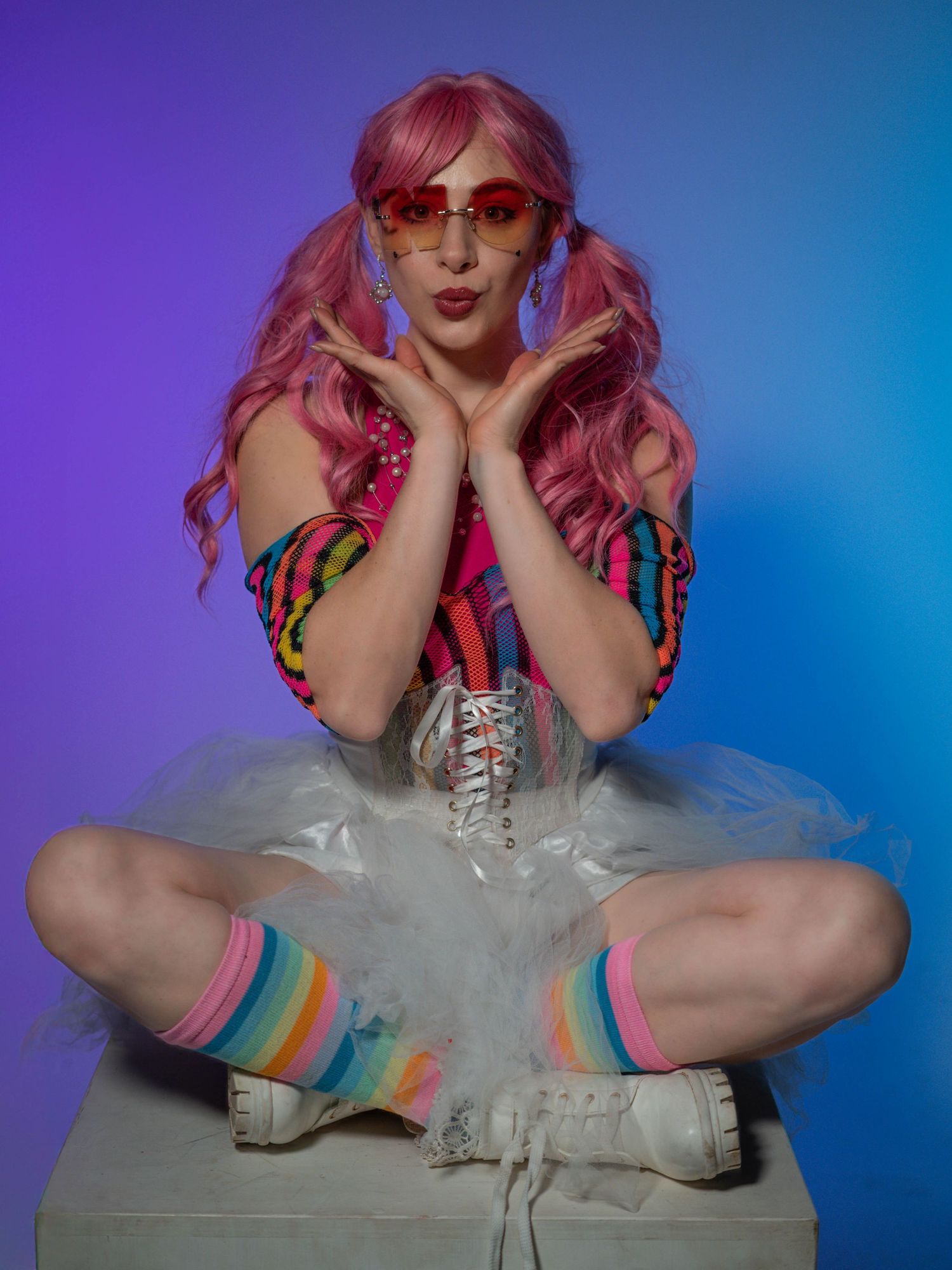 A woman with pink pigtail wig, rainbow socks, white tutu, and red heart sunglasses poses on a box.