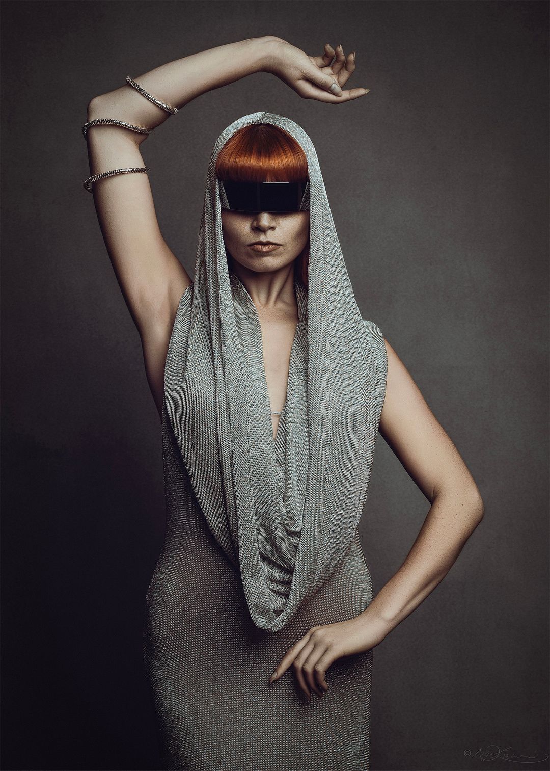 A woman in a gray draped hood and dress with red hair and black visor poses dramatically against a dark background.