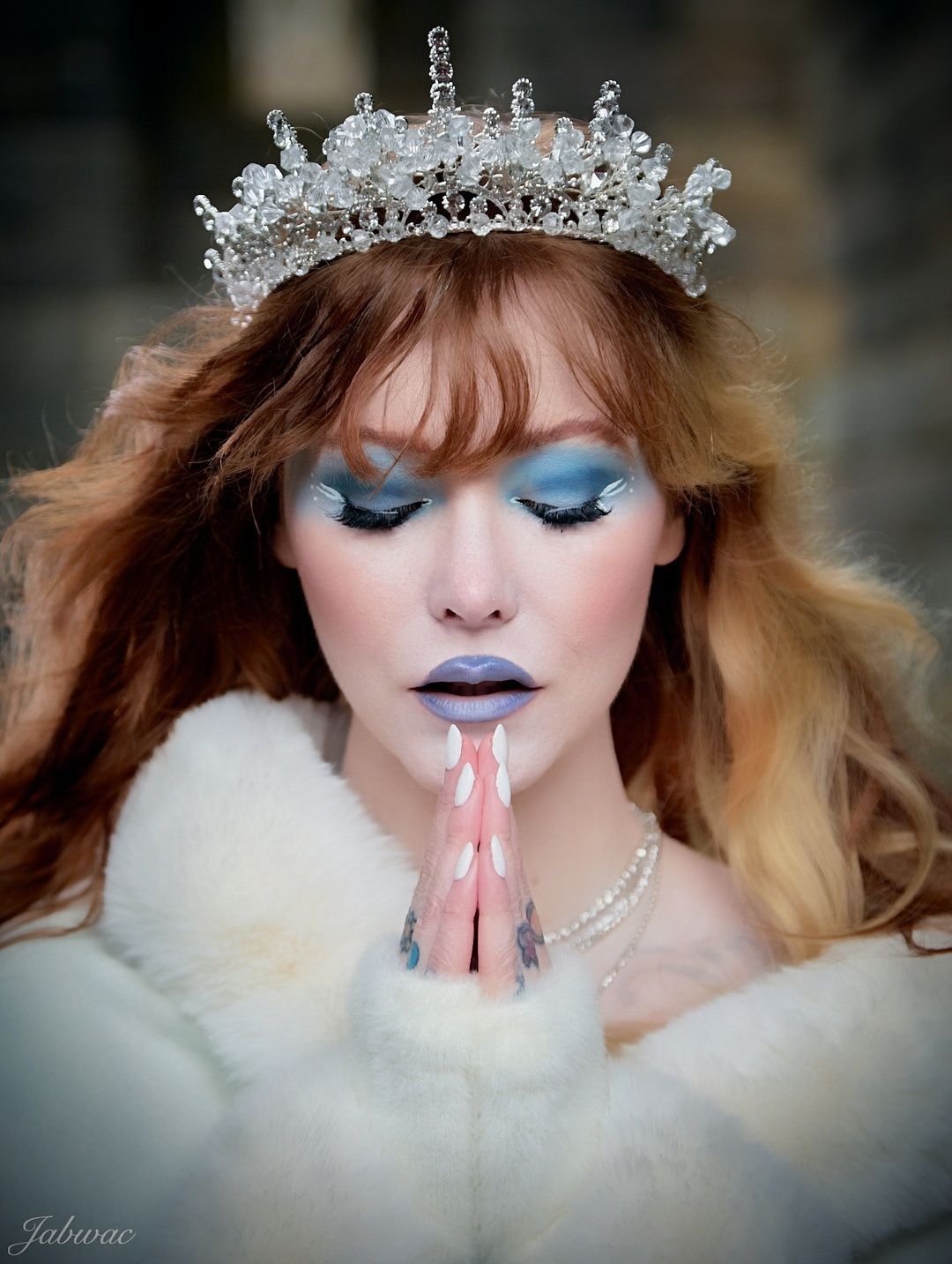 A woman in a crystal tiara and white fur with blue eyeshadow and purple lips poses with hands in prayer.