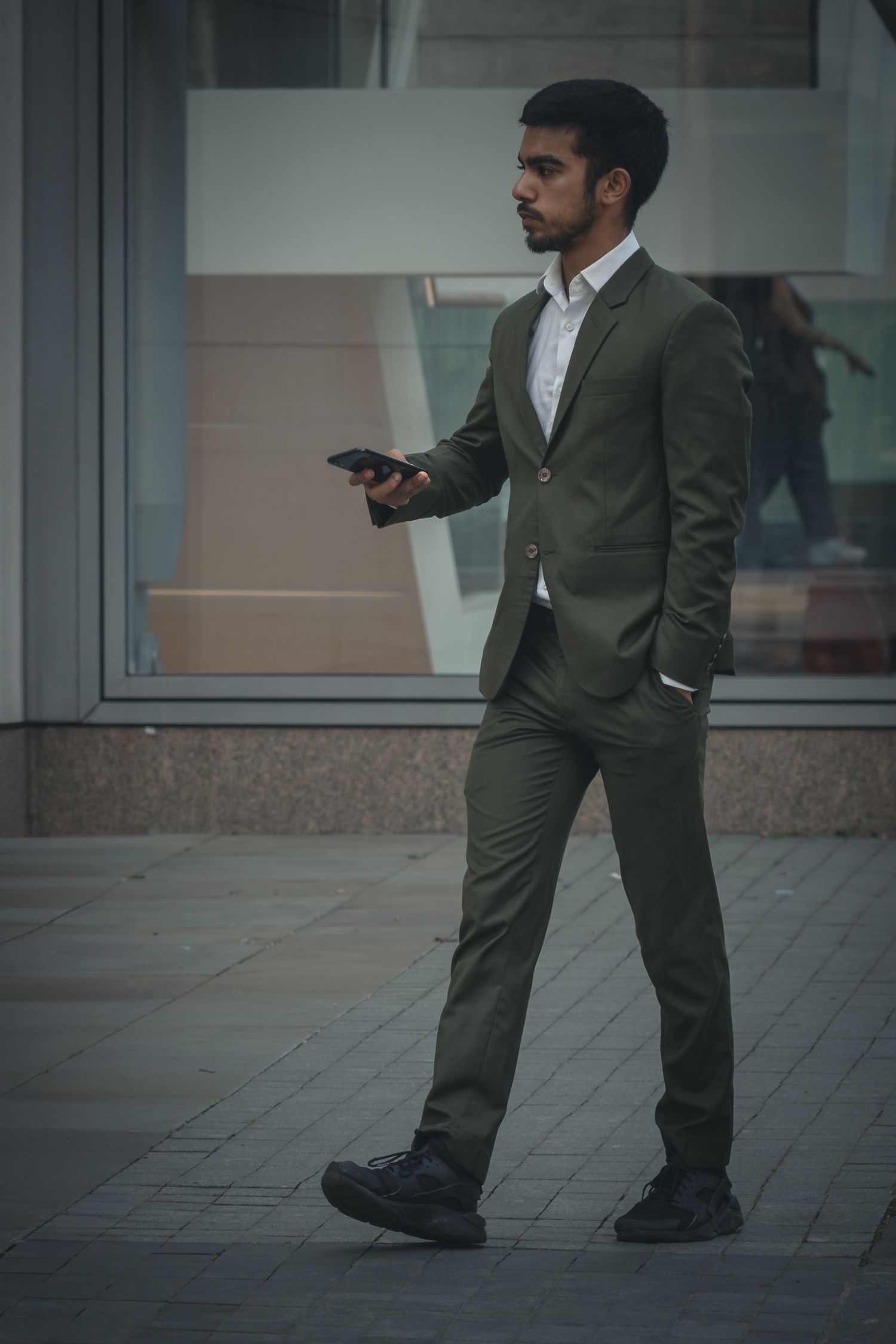A businessman in a dark green suit walks on a paved surface while looking at his mobile phone.