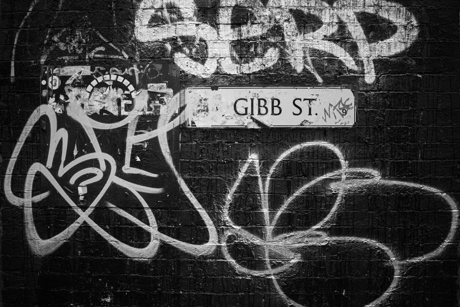 Graffiti tags and street art cover a black wall near Gibb Street, creating an urban street art atmosphere.
