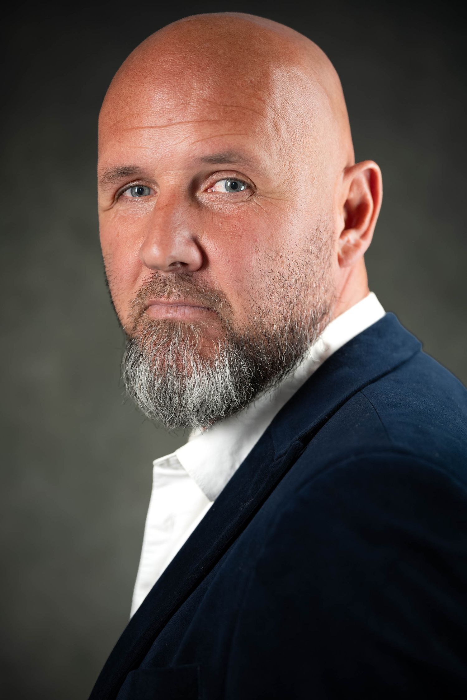 Bald middle-aged man with salt-and-pepper beard wearing navy blazer and white shirt, studio portrait.