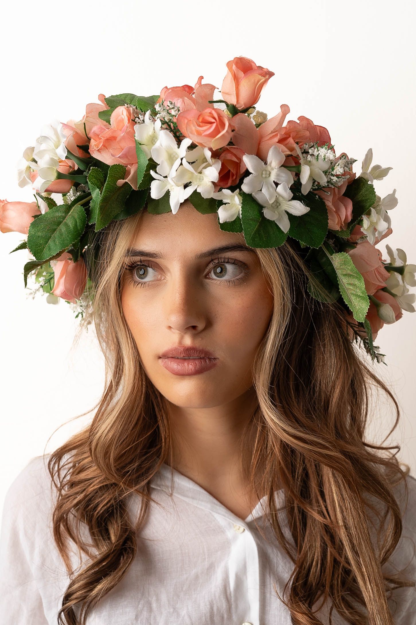Young woman wearing a floral crown of pink roses and white flowers, with long wavy hair and a white top.