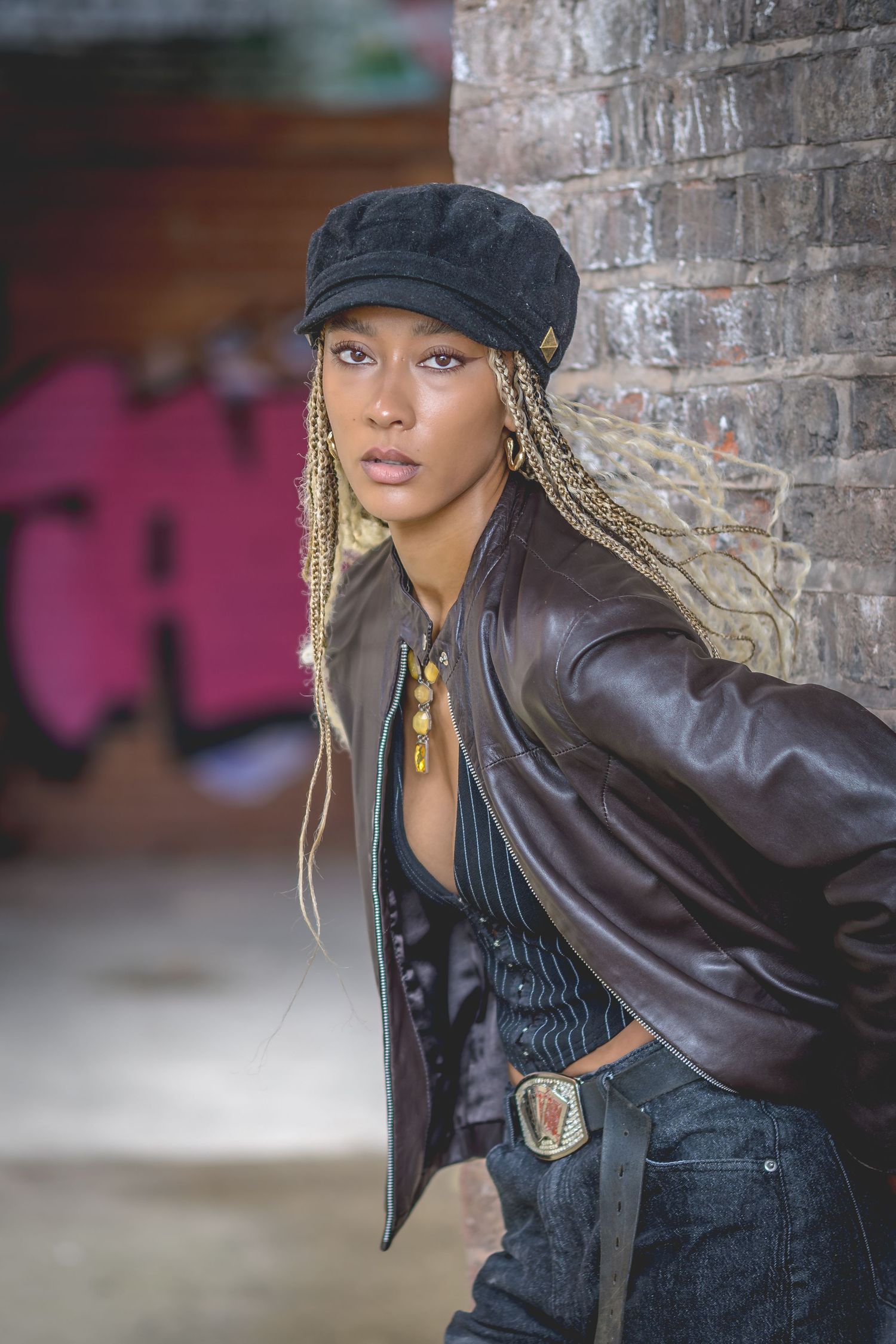 Young woman with blonde braids and black cap leans against brick wall wearing leather jacket and jeans.