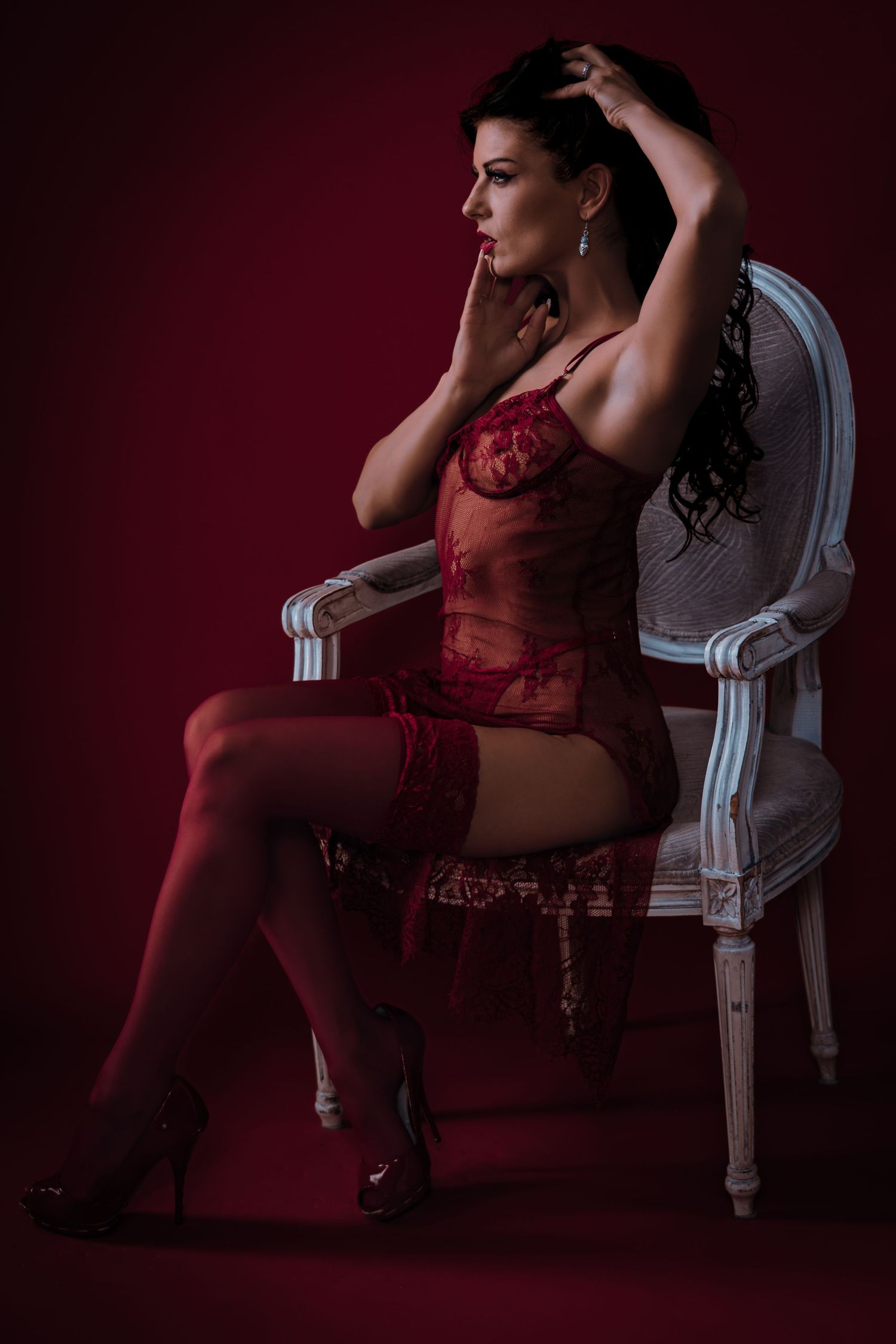 Woman in red lace lingerie and stockings posing elegantly on a white chair against a deep red background.