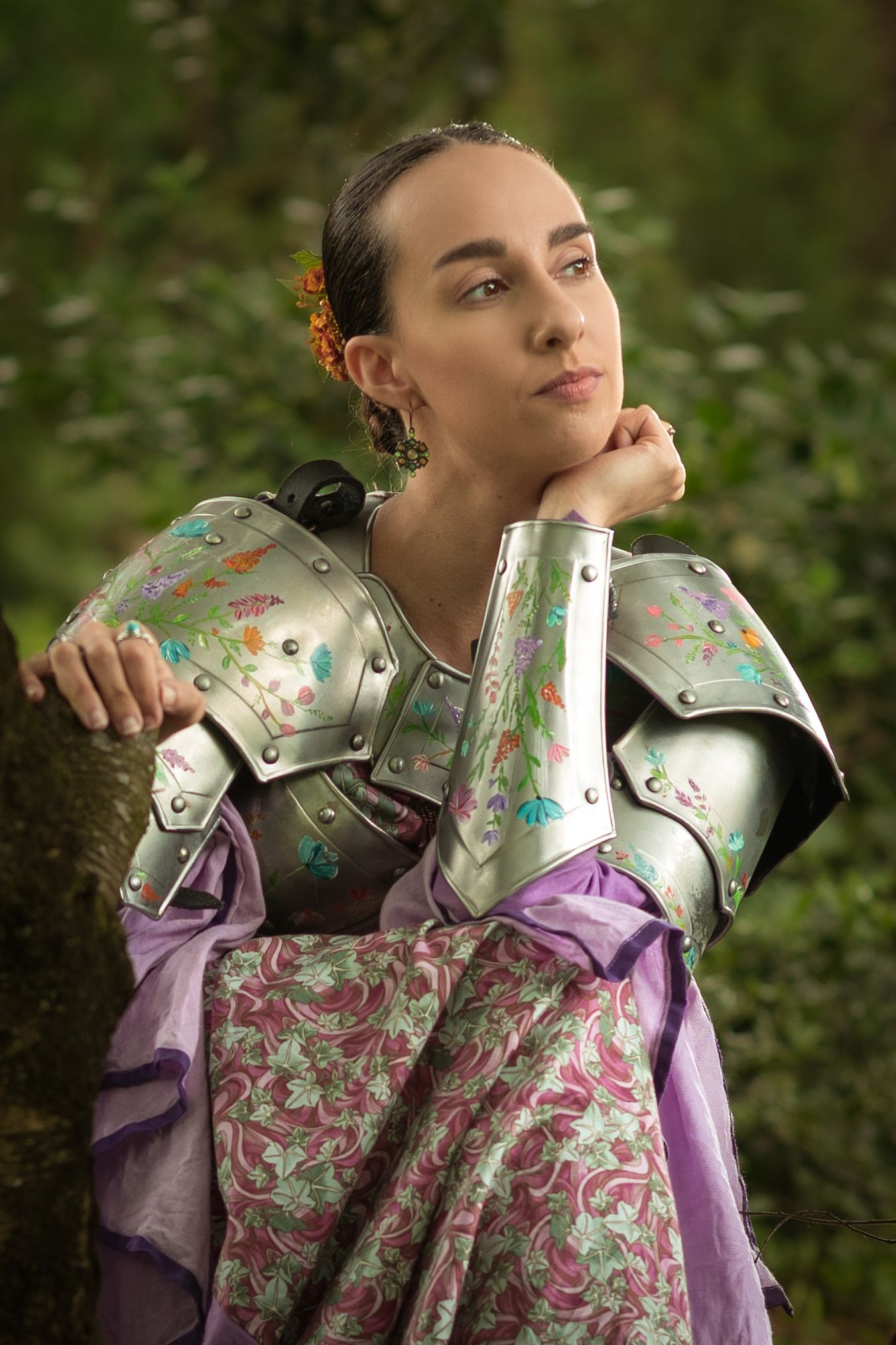 Woman in floral-decorated silver knight armor and purple gown poses thoughtfully in a lush green forest setting.