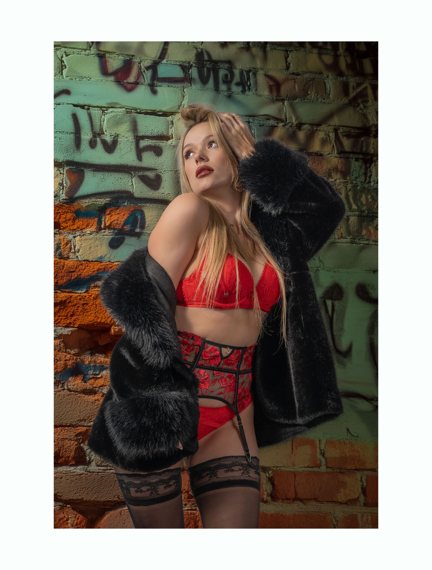 Fashion model in red lingerie and black fur coat poses dramatically against graffiti wall in urban photoshoot setting.