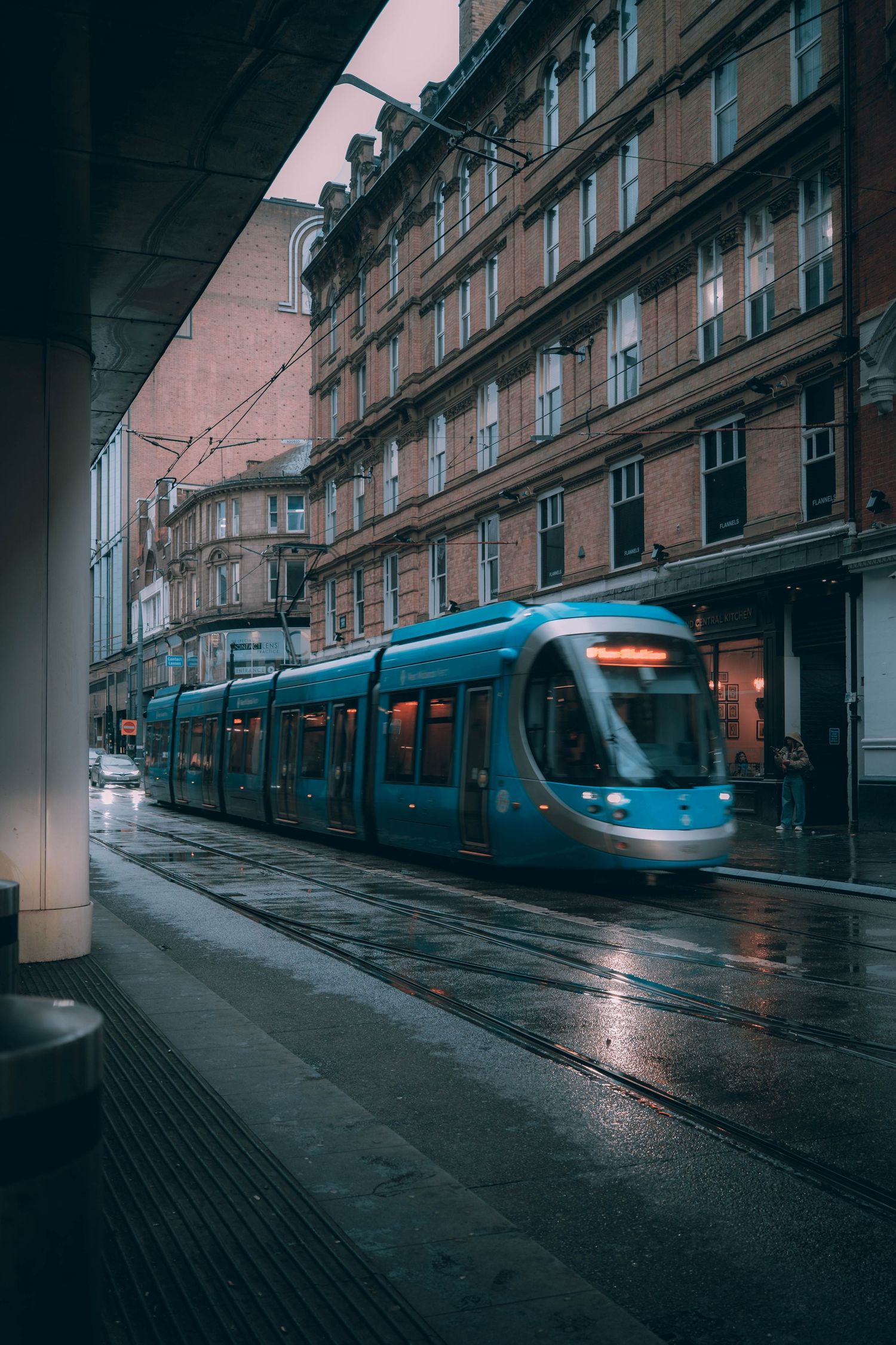 A modern blue tram travels down a wet street in front of historic brownstone buildings on a rainy day.