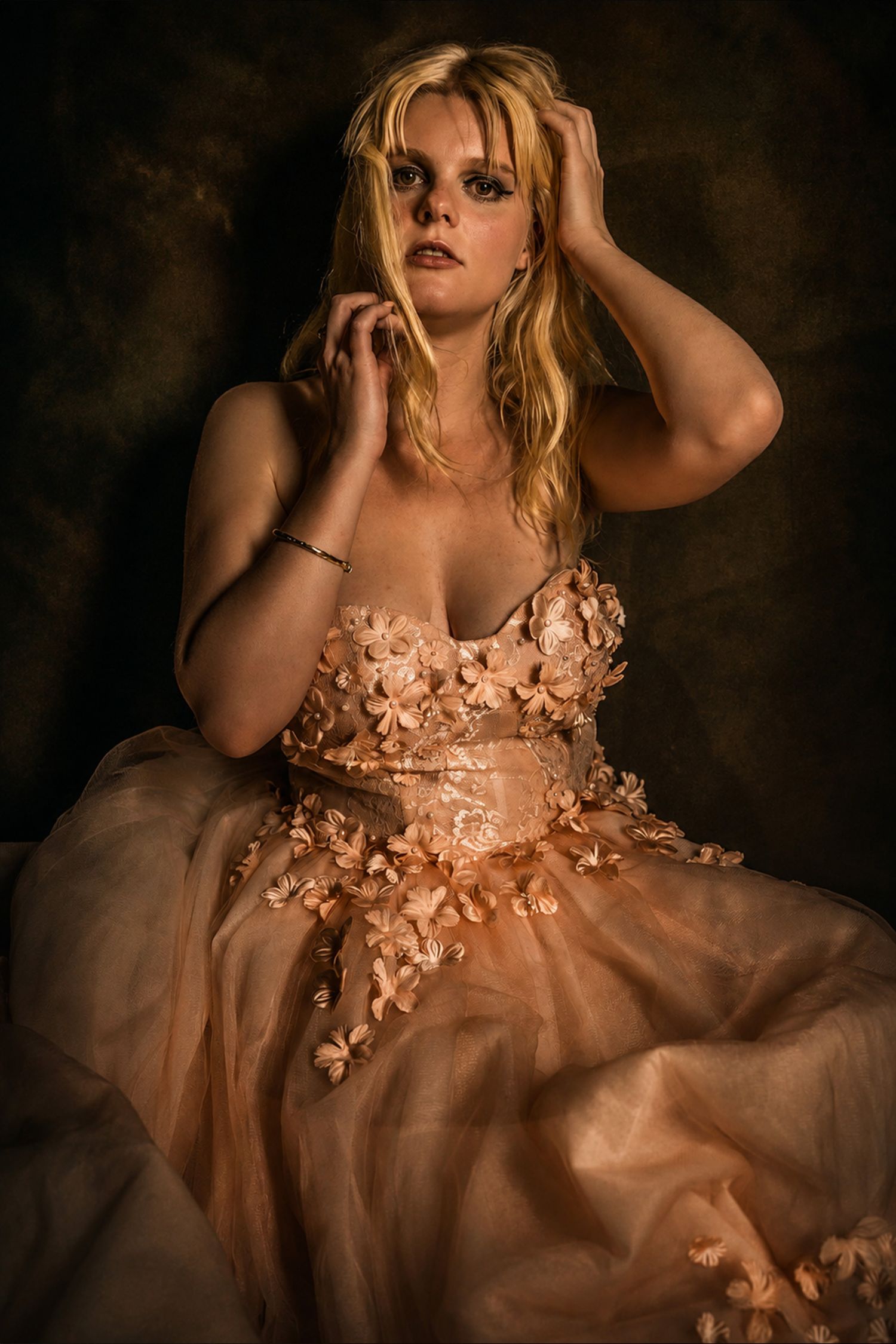 A blonde woman poses in a floral peach ballgown against a dark backdrop, hands raised elegantly in her hair.