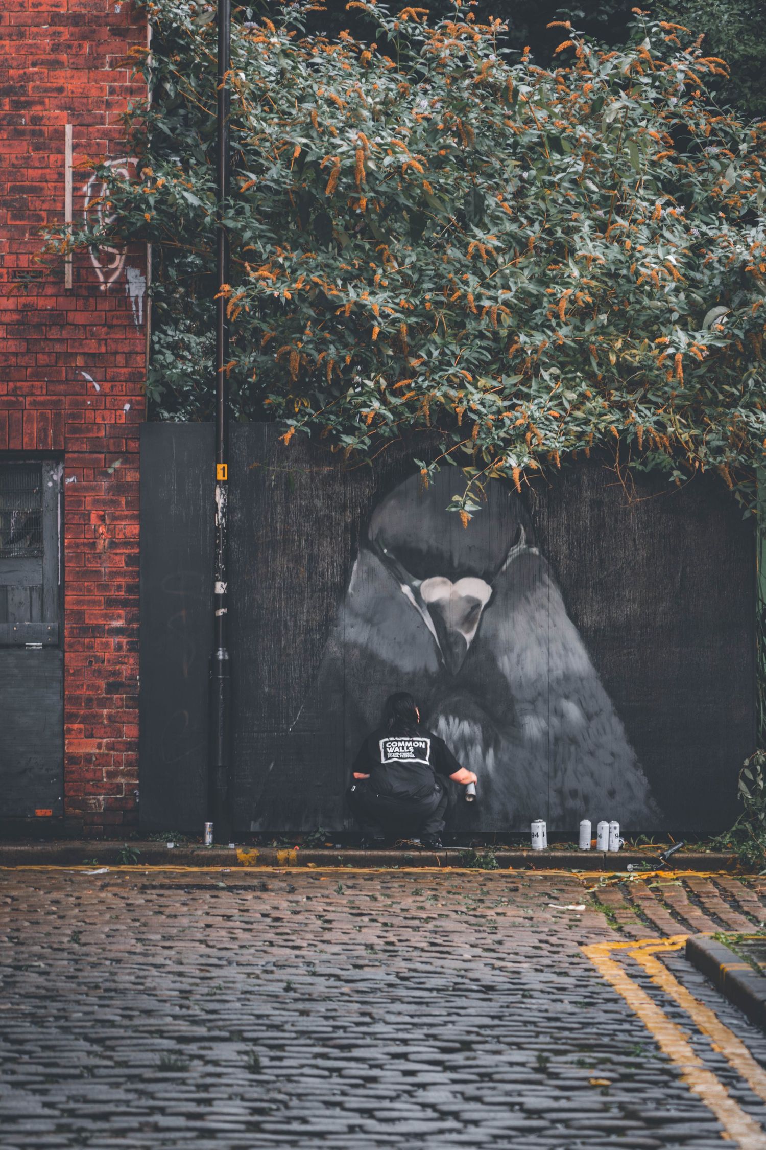 Moody urban street scene with red brick wall, overhanging tree branches, and double yellow lines on wet cobblestone road.