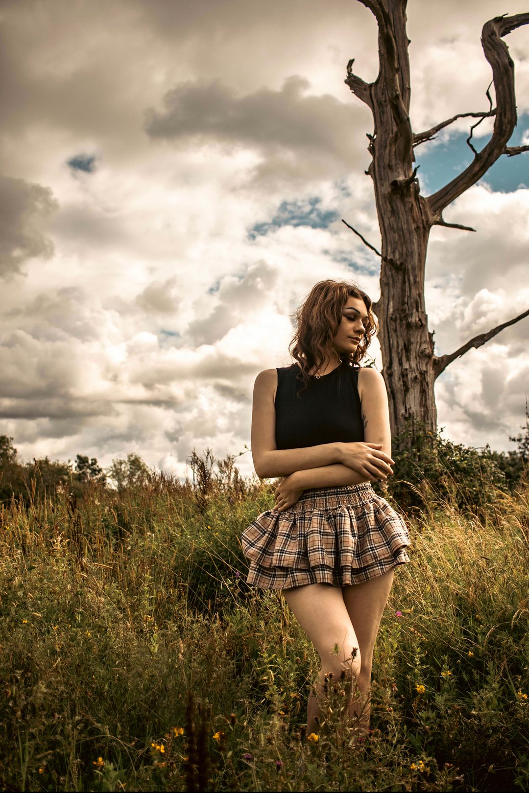 Young woman in plaid mini skirt and black top posing in a wild field near a dead tree under dramatic cloudy sky.
