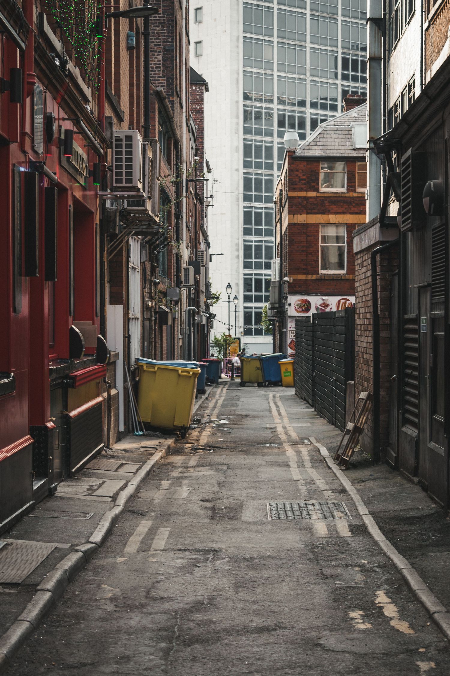 Narrow urban alley between brick buildings with metal pipes and trash bins along the walls.