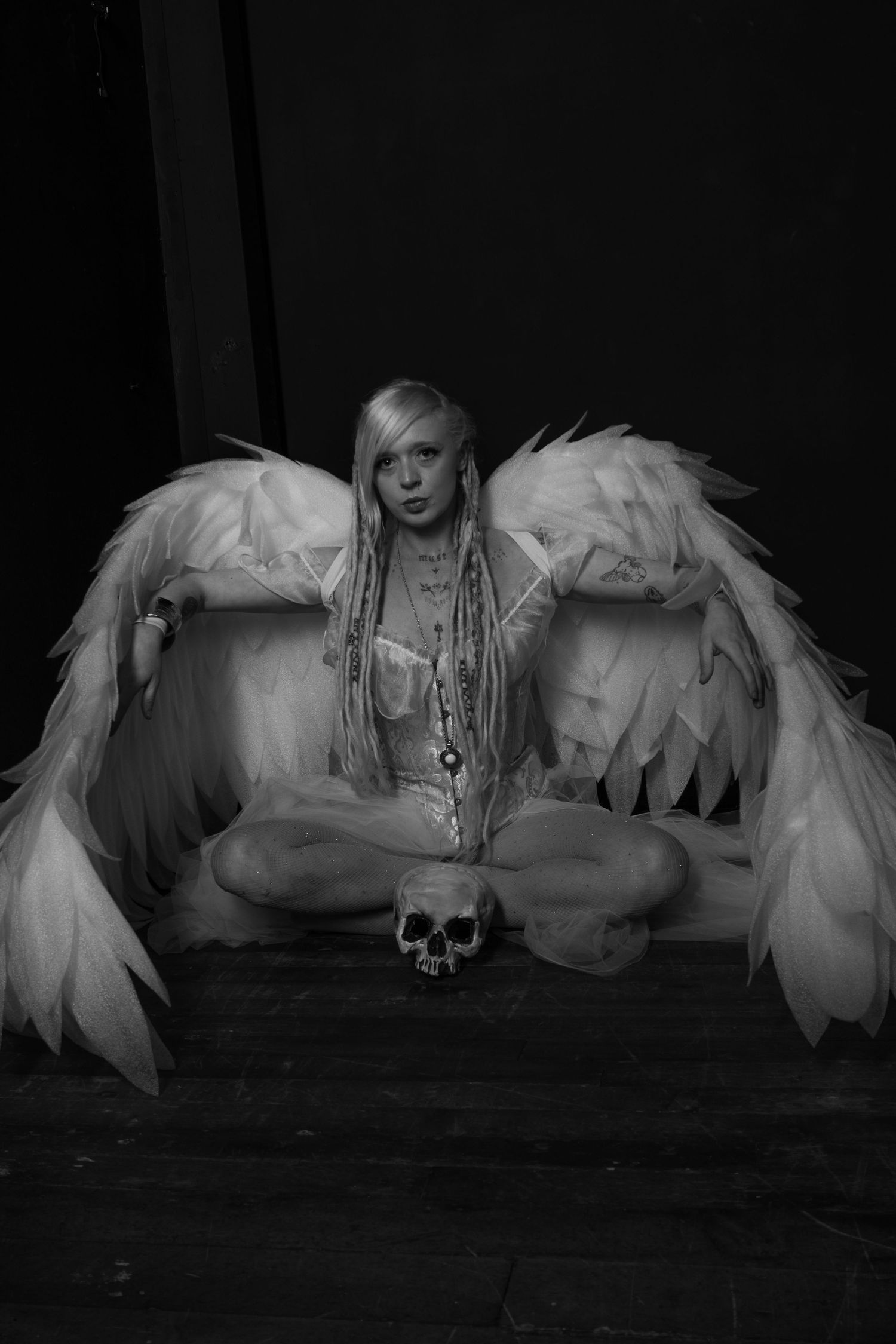 Artistic black and white photo of a figure with white wings sitting cross-legged with a skull, creating a dark gothic aesthetic.