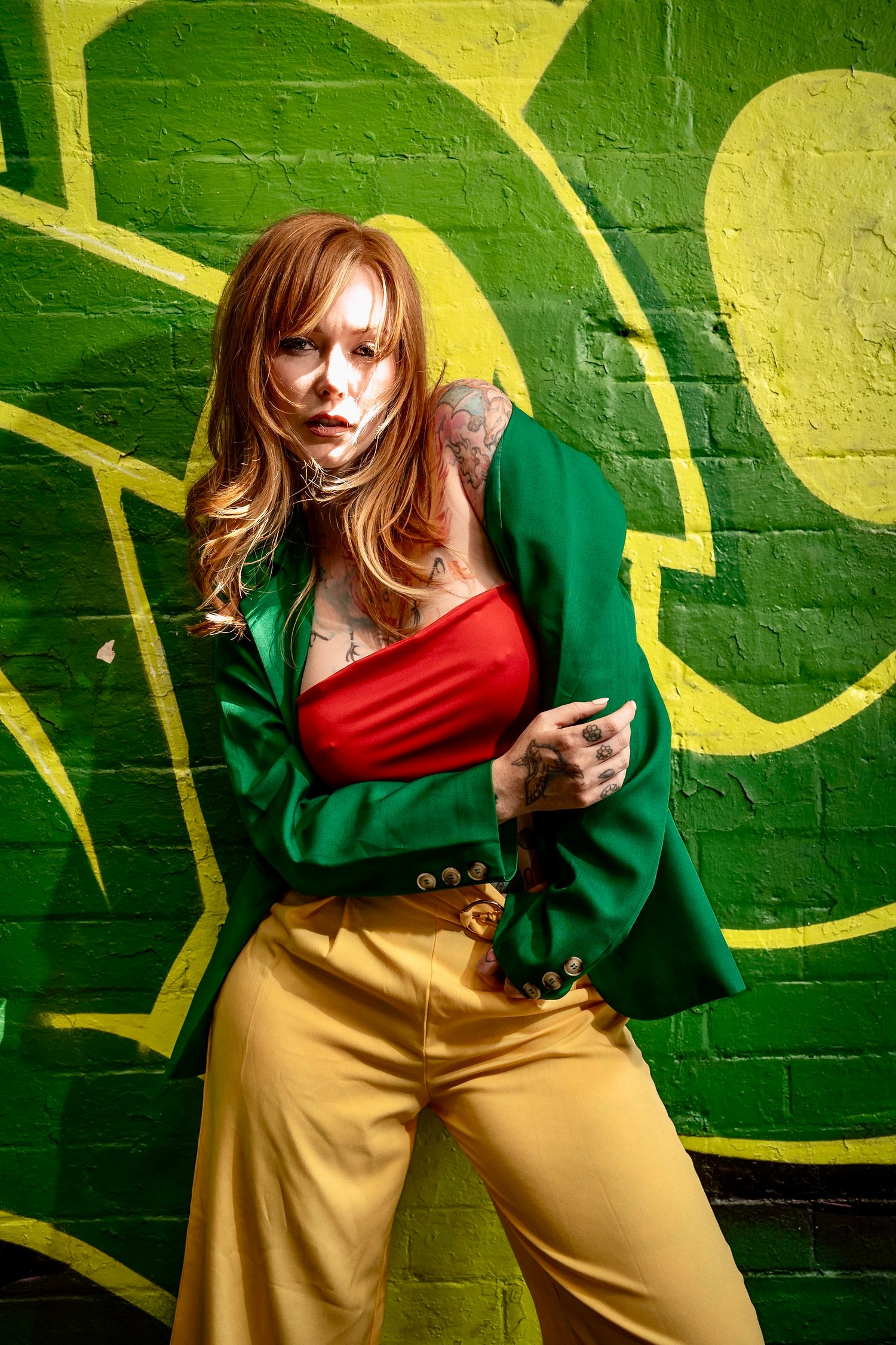 Woman in green blazer, red crop top, and yellow pants posing against a vibrant green graffiti brick wall.