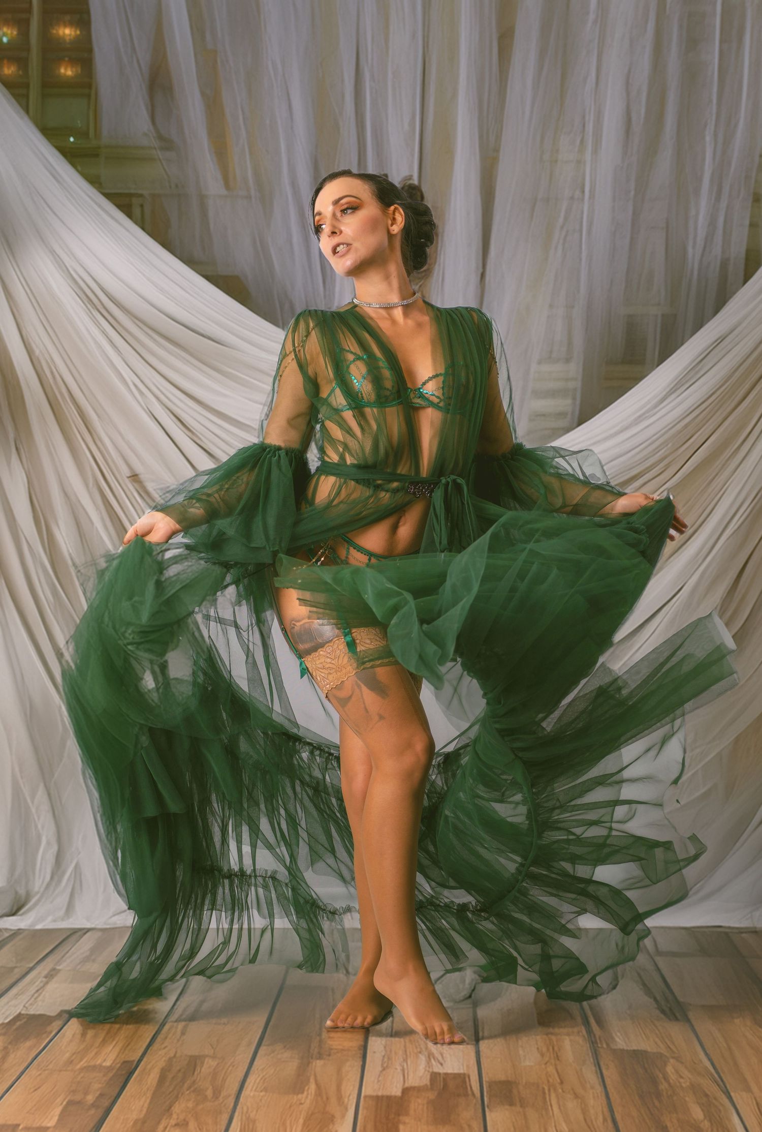 Elegant dancer in flowing emerald green tulle gown poses gracefully against white draped backdrop on wooden floor.