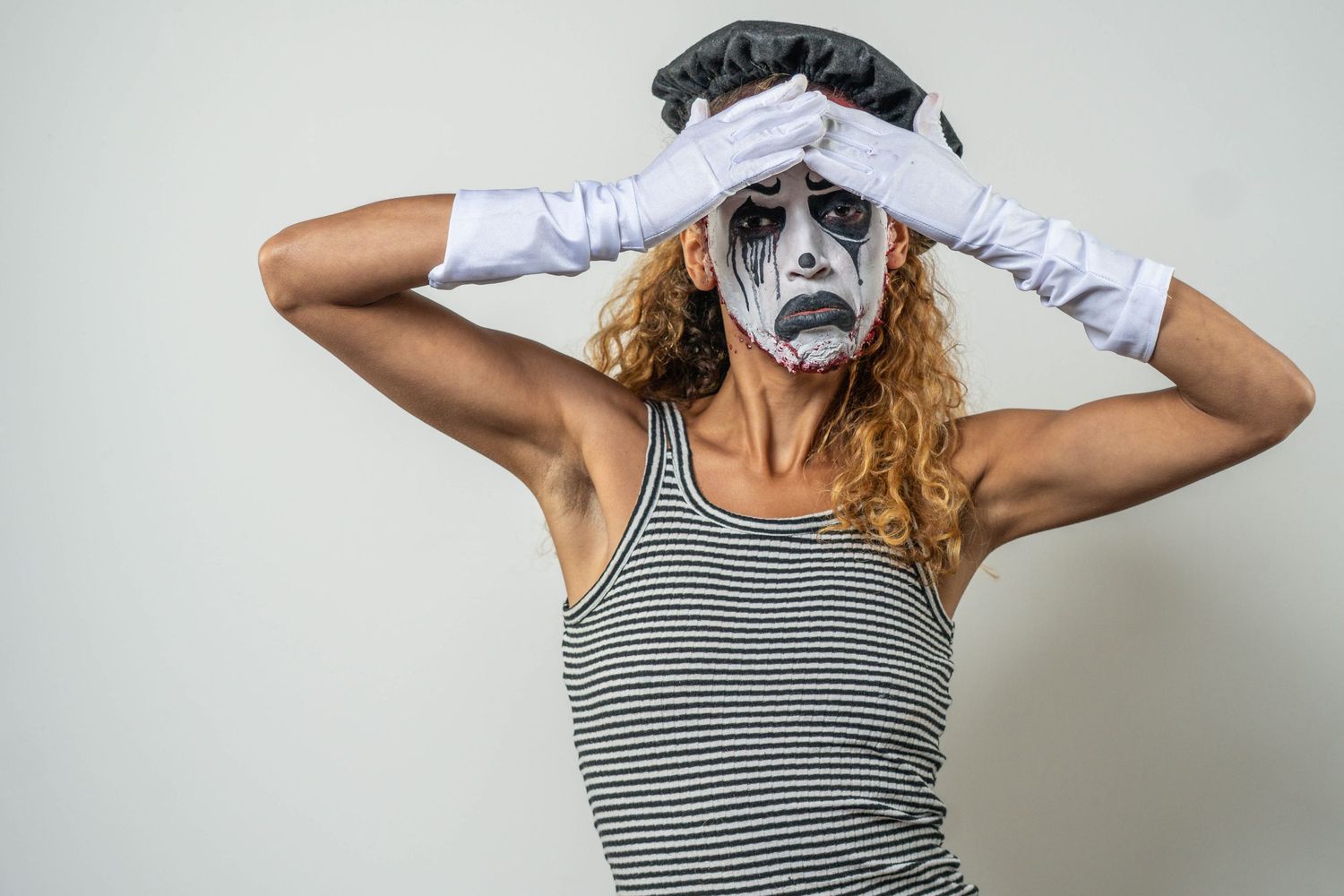 Person in mime-style face paint and striped tank top poses with hands on head against light background.