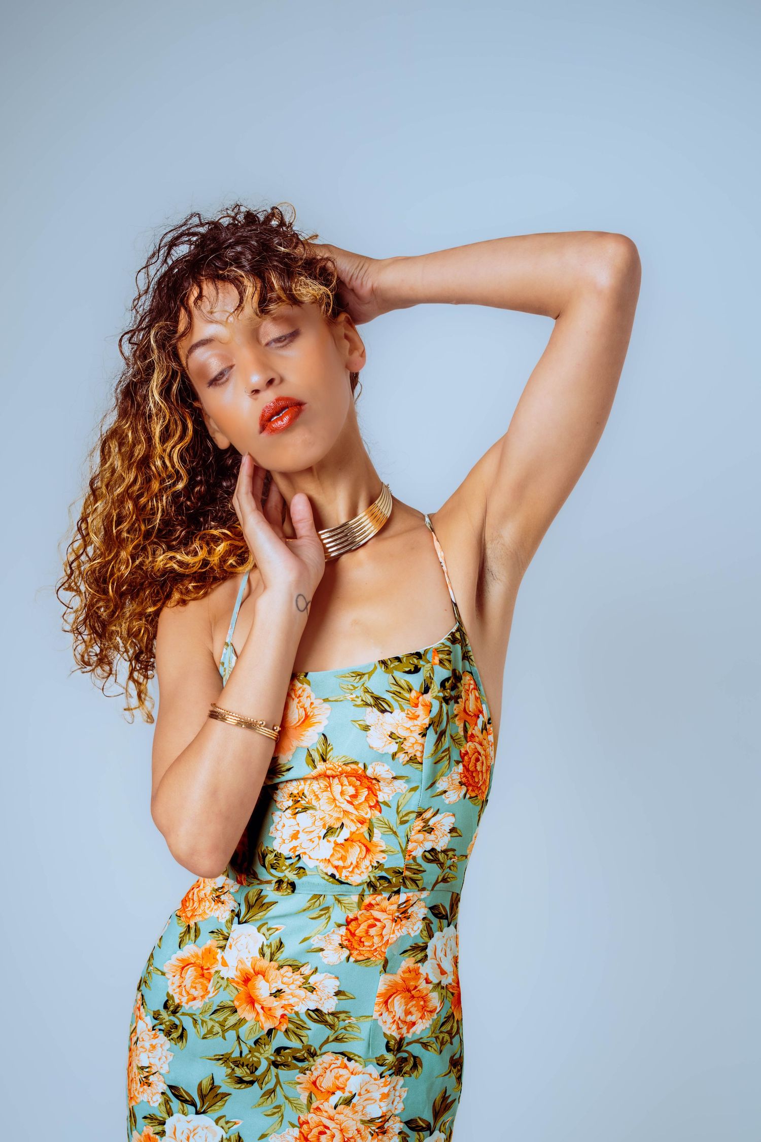 Woman in floral dress with curly hair, gold choker, posing gracefully against a light blue background.