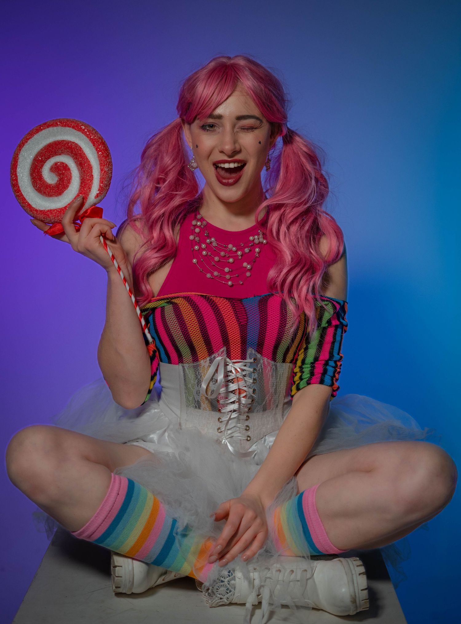 A woman in pink wig, rainbow striped top, tutu skirt, and colorful socks holds a large lollipop against purple background.
