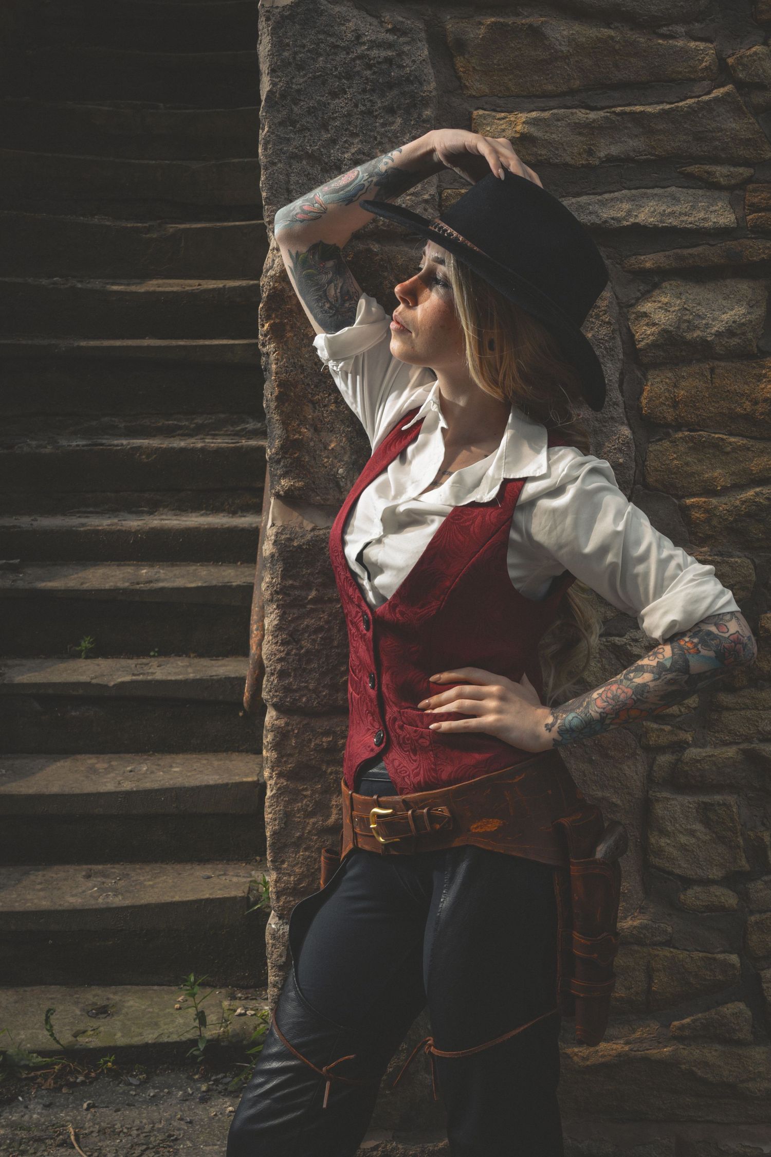Tattooed woman in cowboy hat, red vest, and white shirt poses against stone wall with stairs.