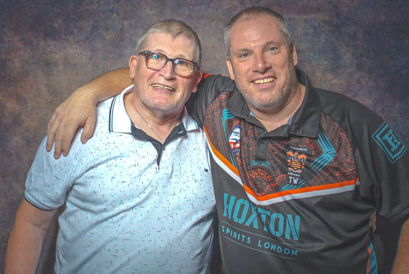 Two smiling men pose together, one wearing glasses and a blue shirt, the other in a Hoxton Spirits London rugby jersey.