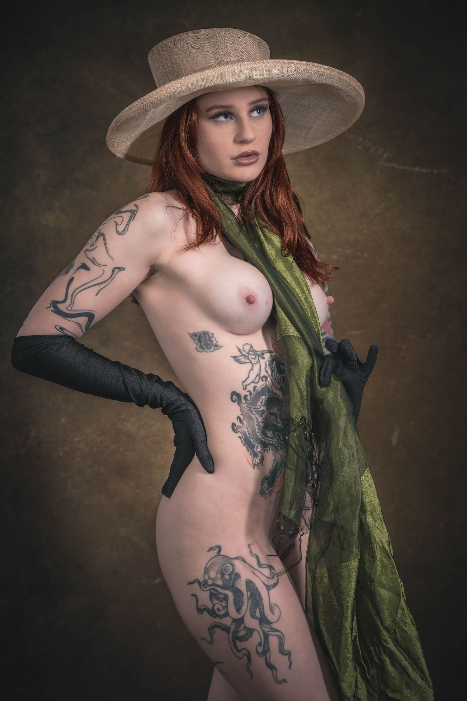 Tattooed redhead woman posing in wide-brim hat, green scarf, and black gloves in artistic fine art portrait.