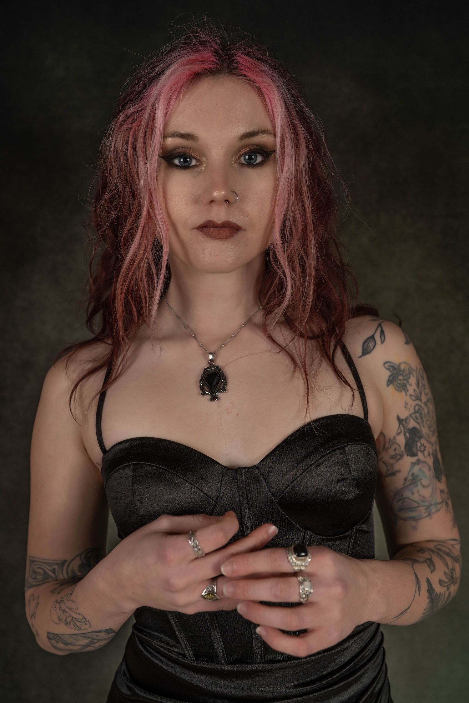 Young woman with pink hair, tattoos, and black corset poses confidently wearing silver rings and a dark pendant necklace.