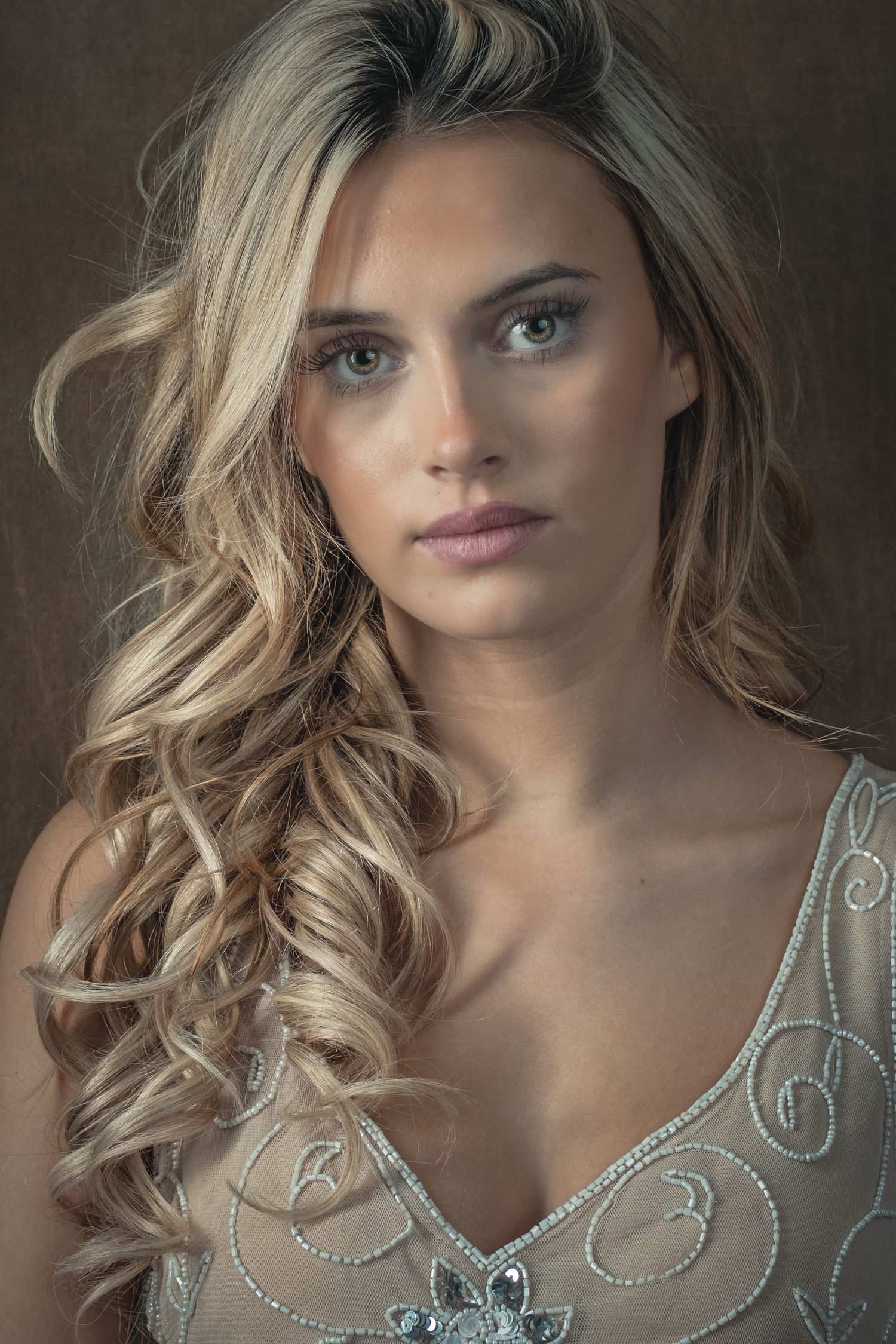 Portrait of a young woman with long blonde curly hair wearing a silver beaded dress against a dark background.