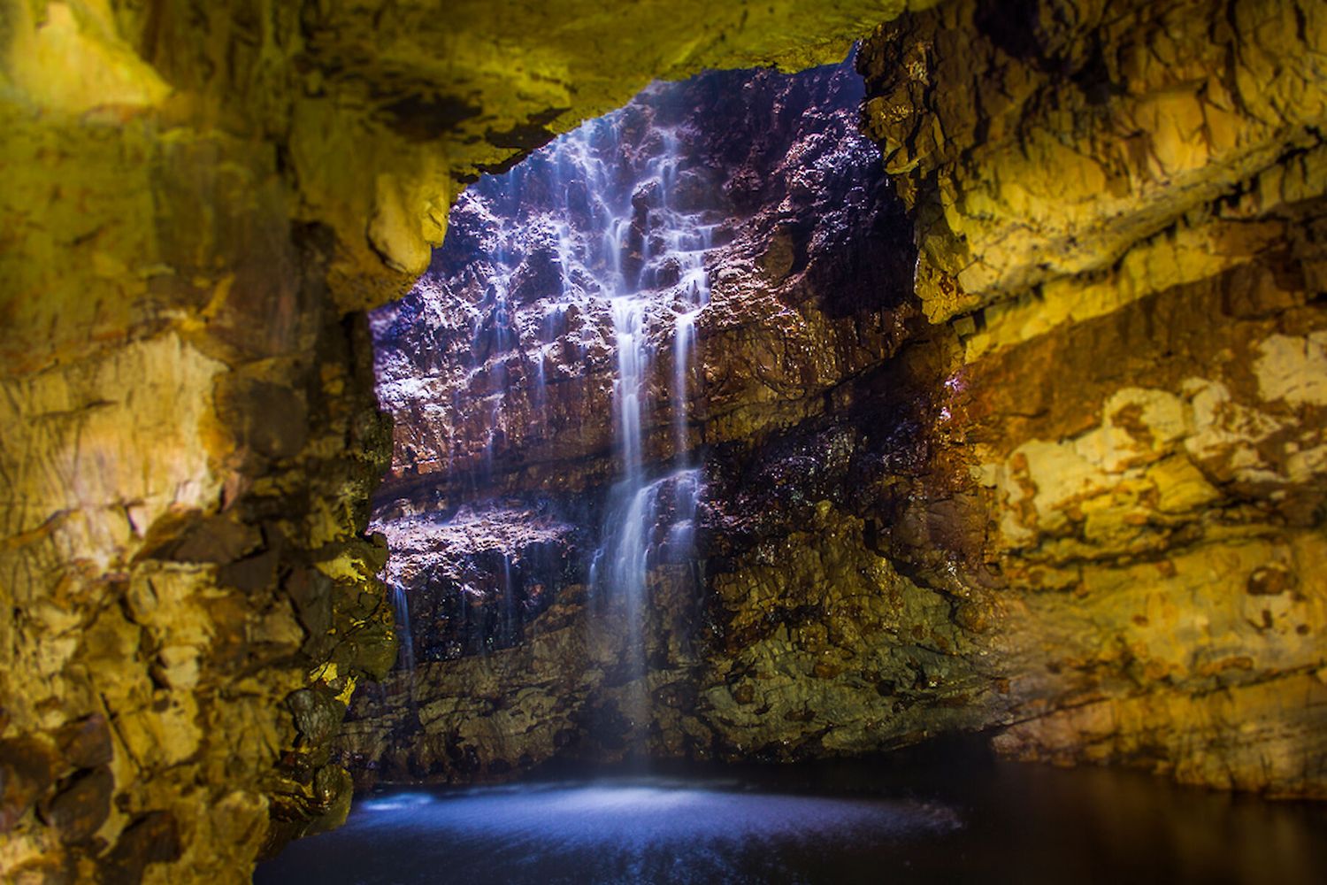 A scenic waterfall flows through a dark limestone cave with dramatic lighting illuminating the rocky formations.