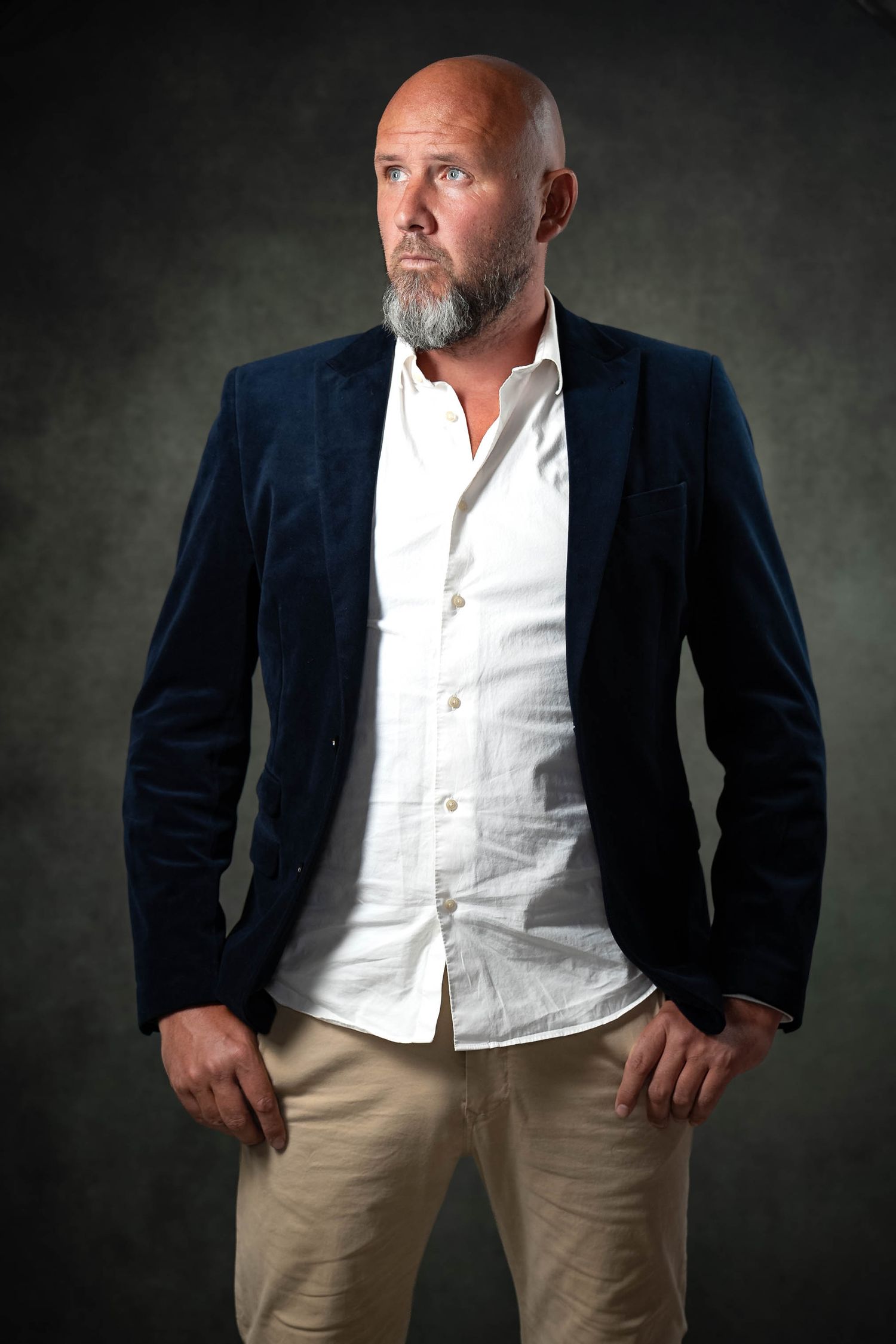 Bald man with gray beard wearing navy blazer and white shirt poses confidently against dark background.