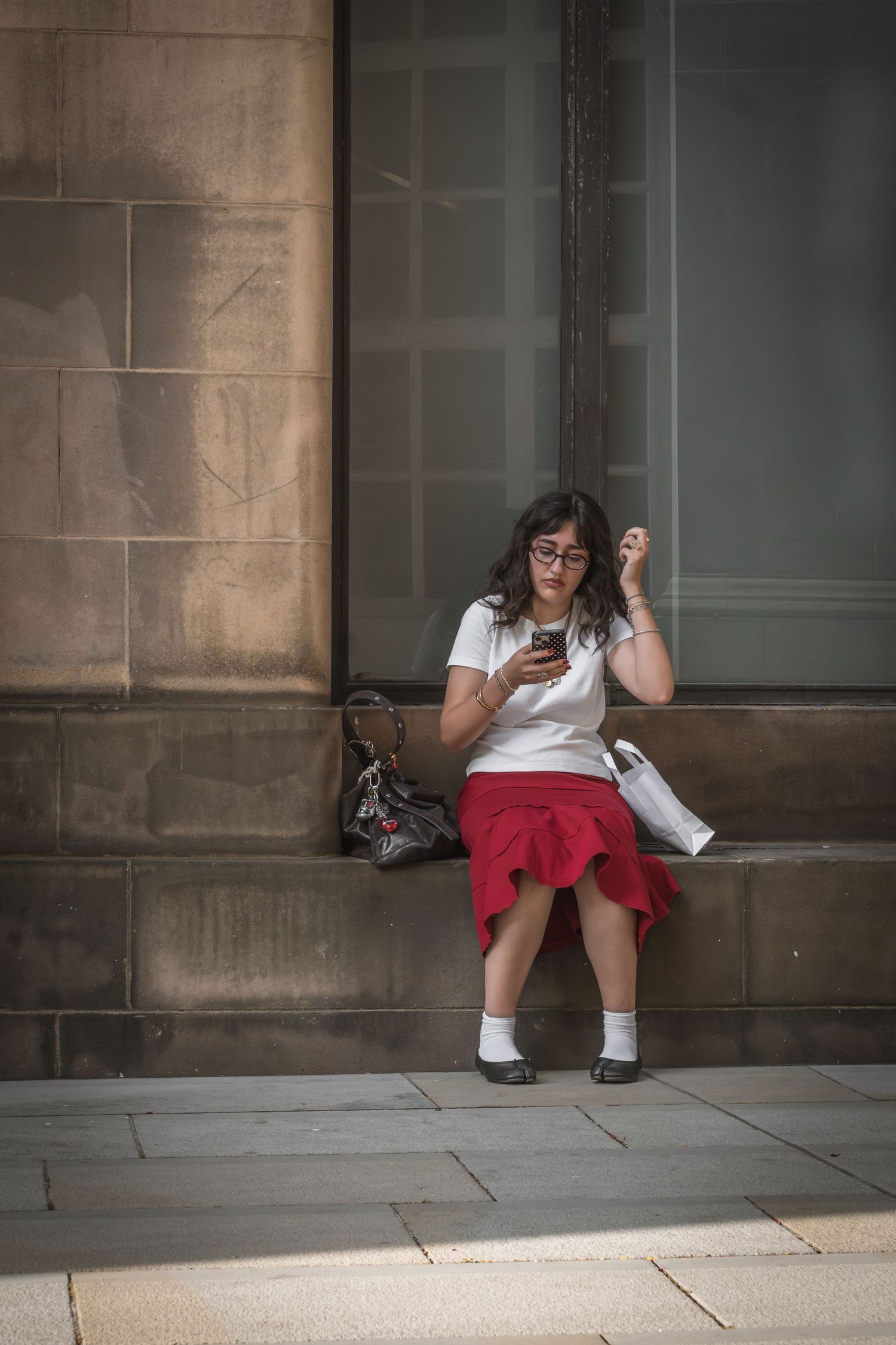 Someone in a white top and red skirt sits on a stone ledge beside a glass building wall.