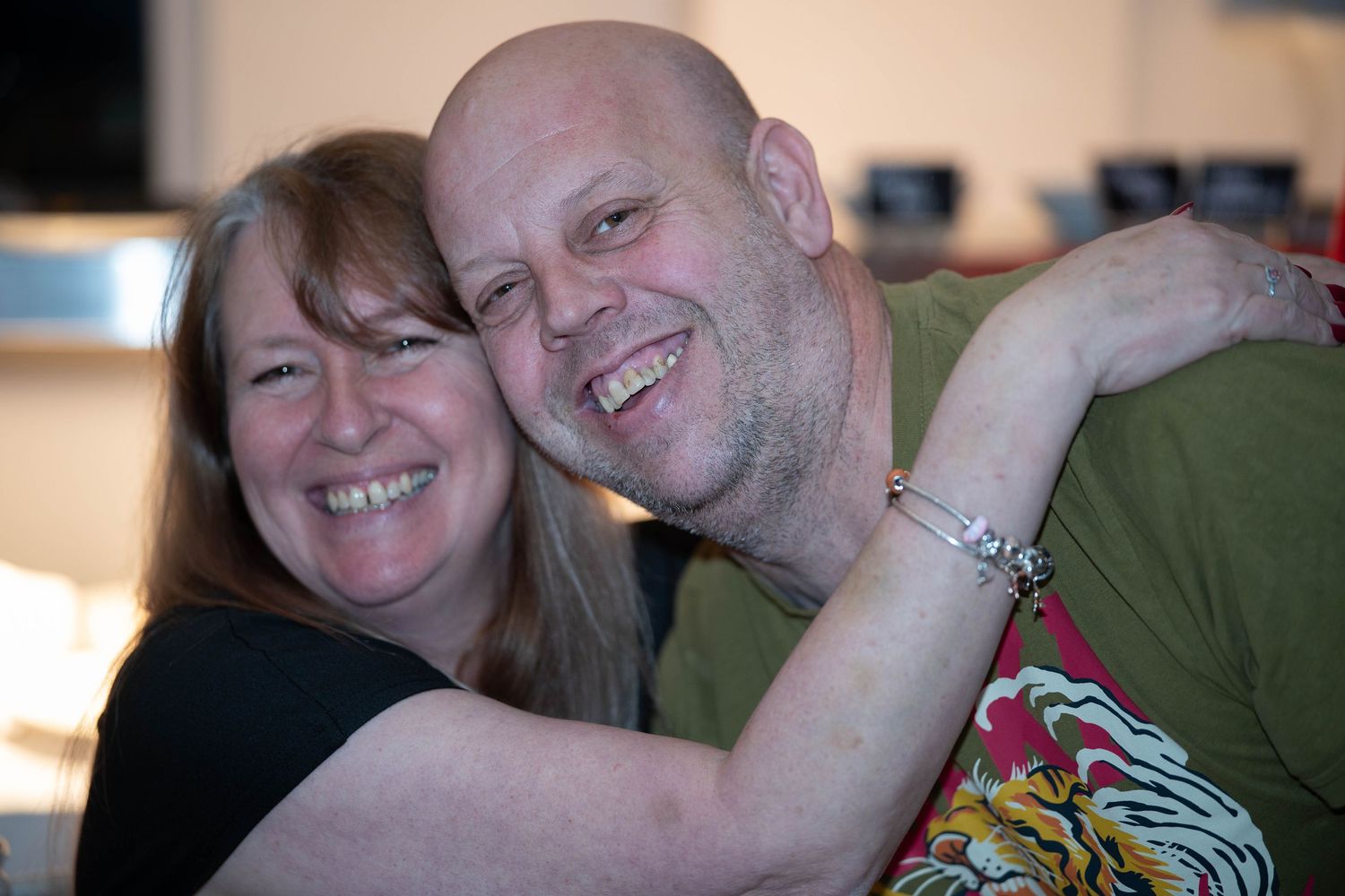 A smiling woman with brown hair hugs a bald man in a green shirt, both laughing warmly indoors.