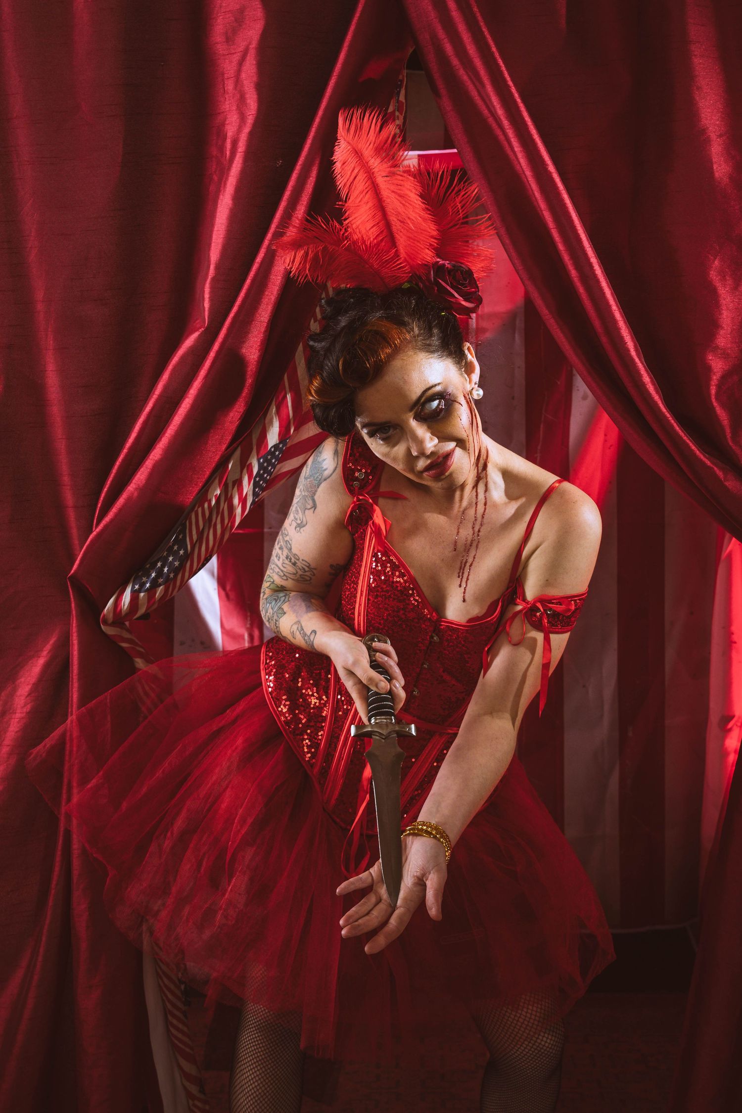 Woman in red sequin corset and tutu with feathered headpiece poses dramatically behind red curtains.