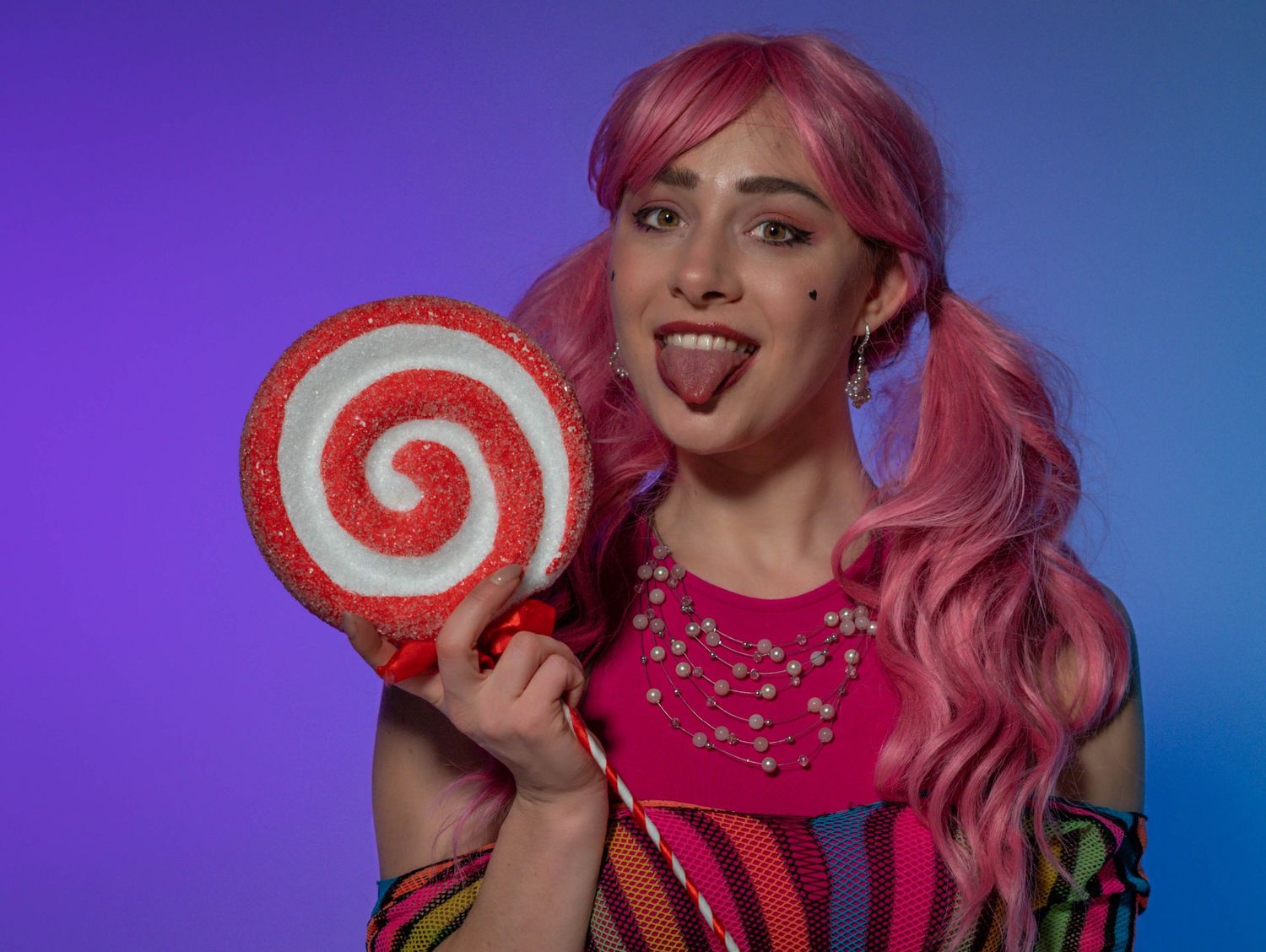 A person with pink hair holds a large red and white swirl lollipop against a purple and blue gradient background.
