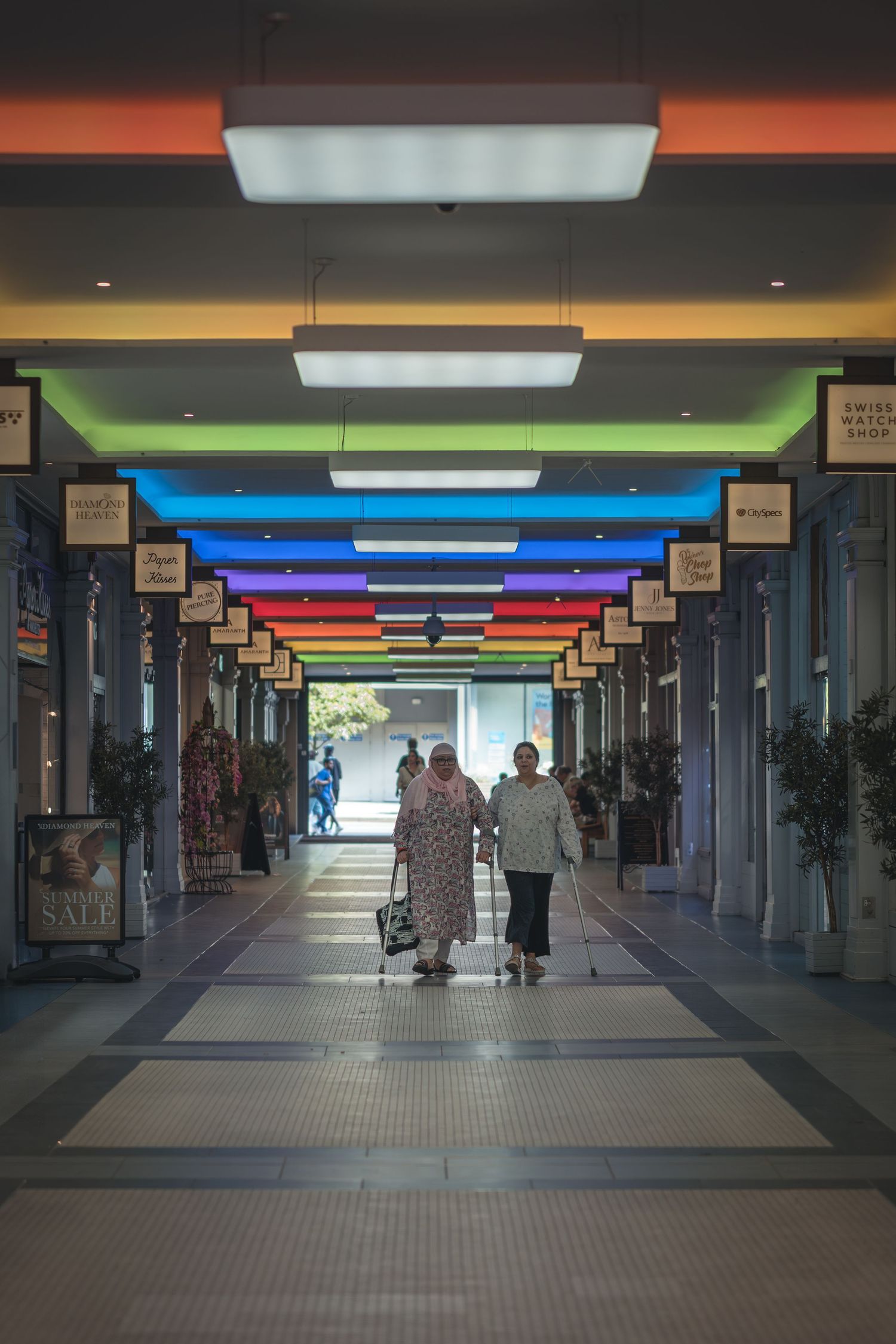 A colorful shopping arcade corridor with rainbow-colored lighting panels on the ceiling creating a gradient effect.
